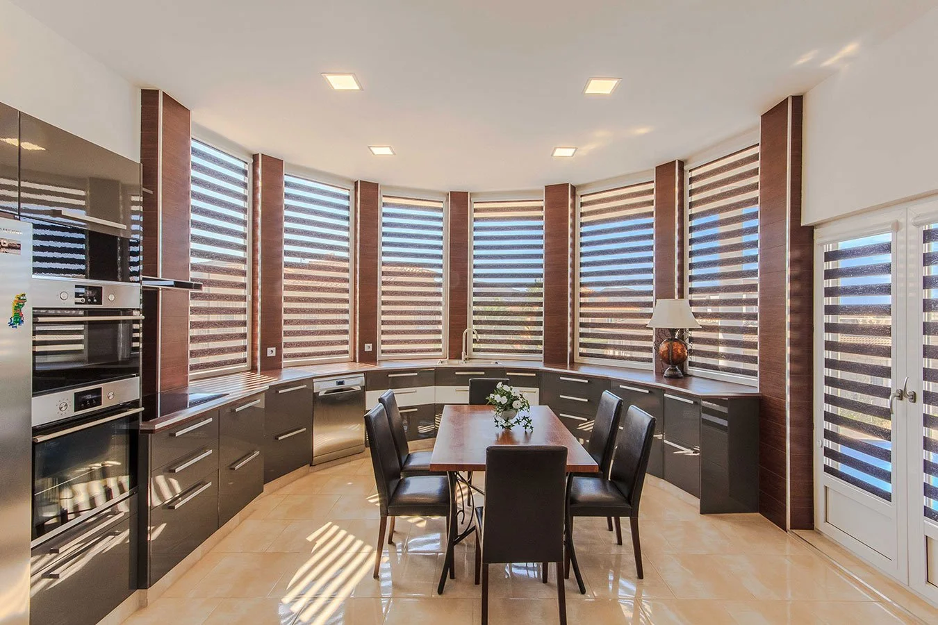Cocina moderna con ventanas grandes con persianas de diseño blanco y negro, mesa de madera con seis sillas negras, cerámica beige en el piso, electrodomésticos de acero inoxidable, lámpara y decoración mínima.