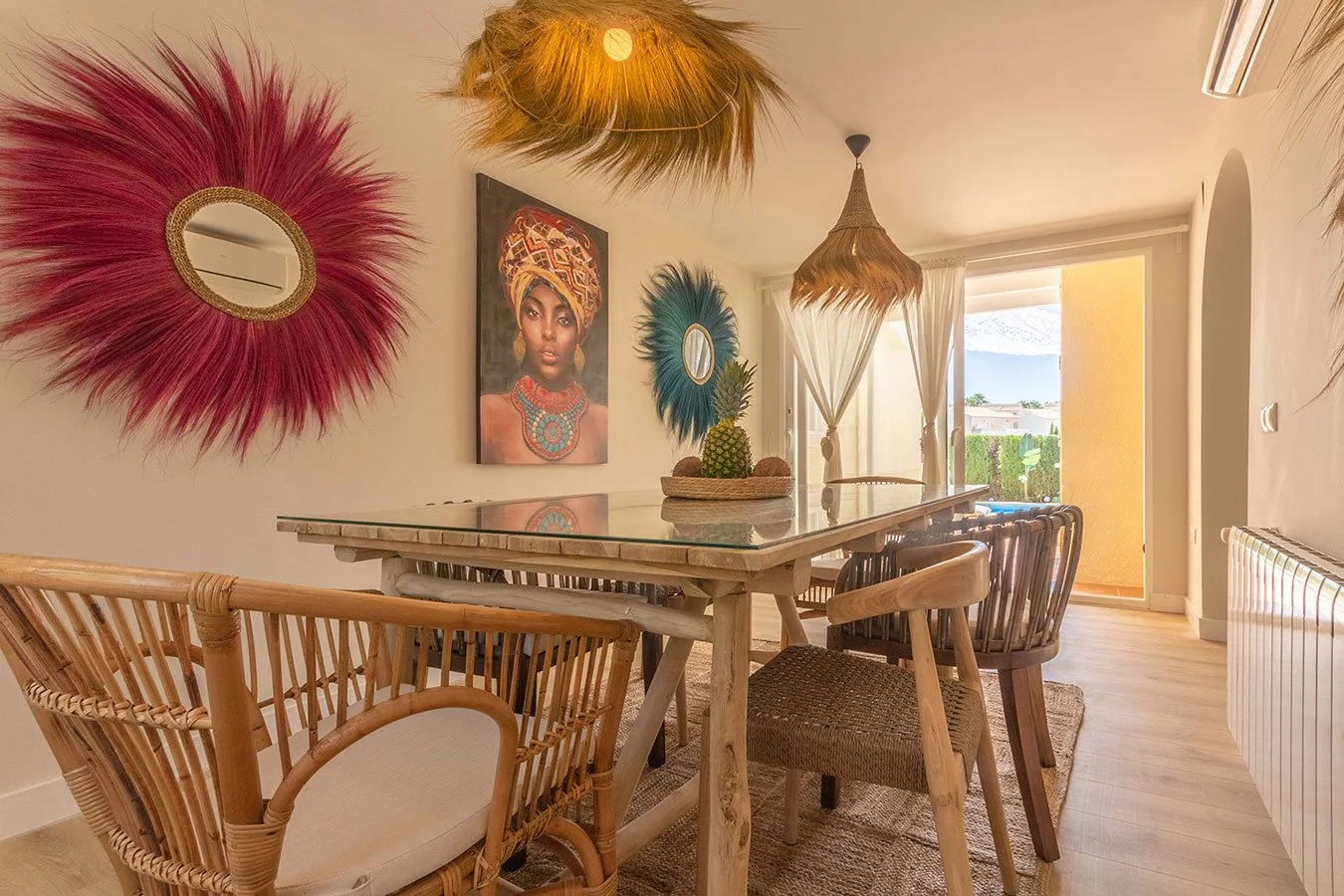 Sala de comedor con decoración bohemia, con espejos con marcos de paja en la pared y una pintura de una mujer africana. Mesa de madera con frutas y plantas en el centro, paredes claras, cortinas blancas y puerta hacia un patio con vista a un cielo az