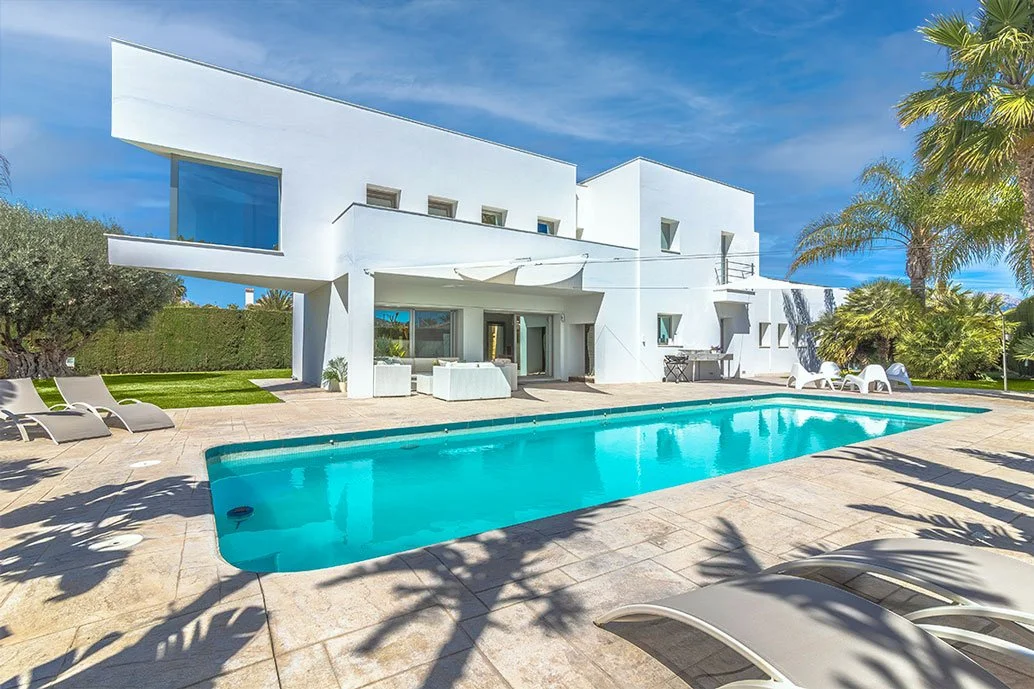 Casa moderna blanca con piscina rectangular y tumbonas en la zona exterior, rodeada de palmeras y vegetación, bajo un cielo despejado.