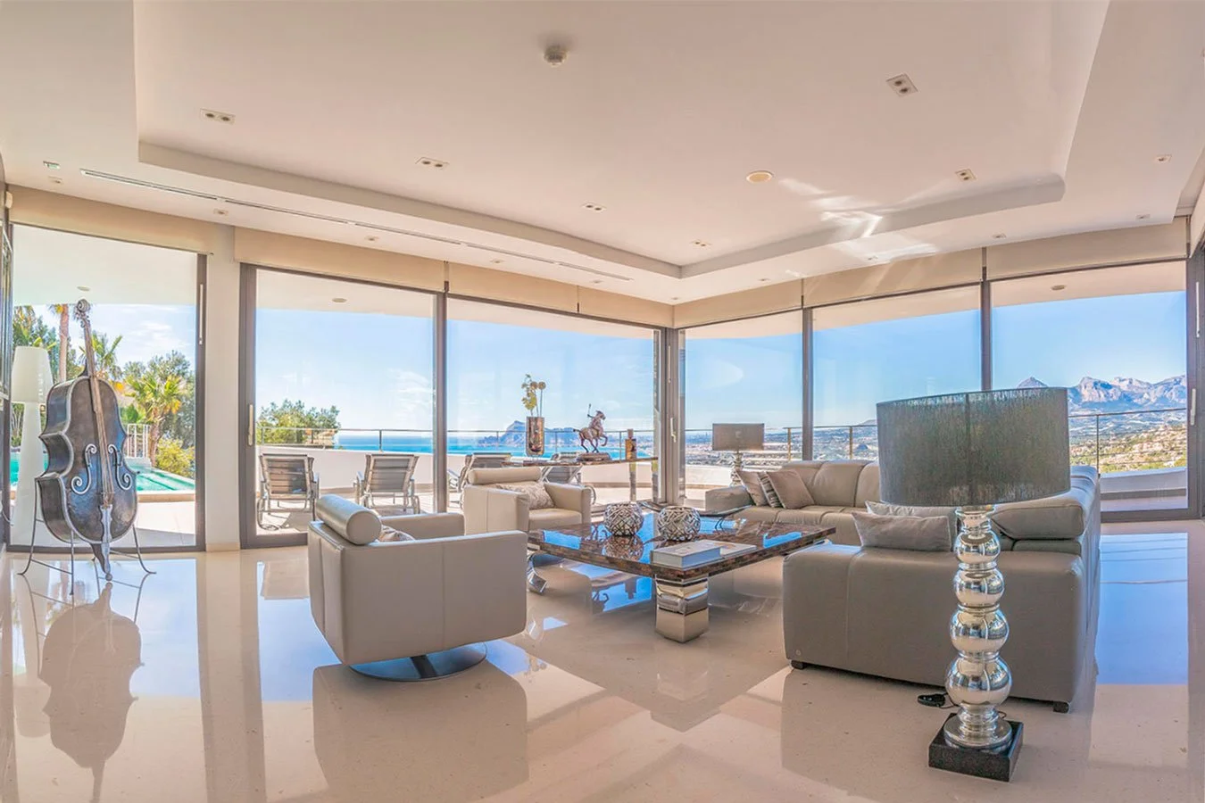 Sala de estar moderna con vista panorámica al mar y montañas, sillones blancos y una mesa de centro de madera con decoraciones.