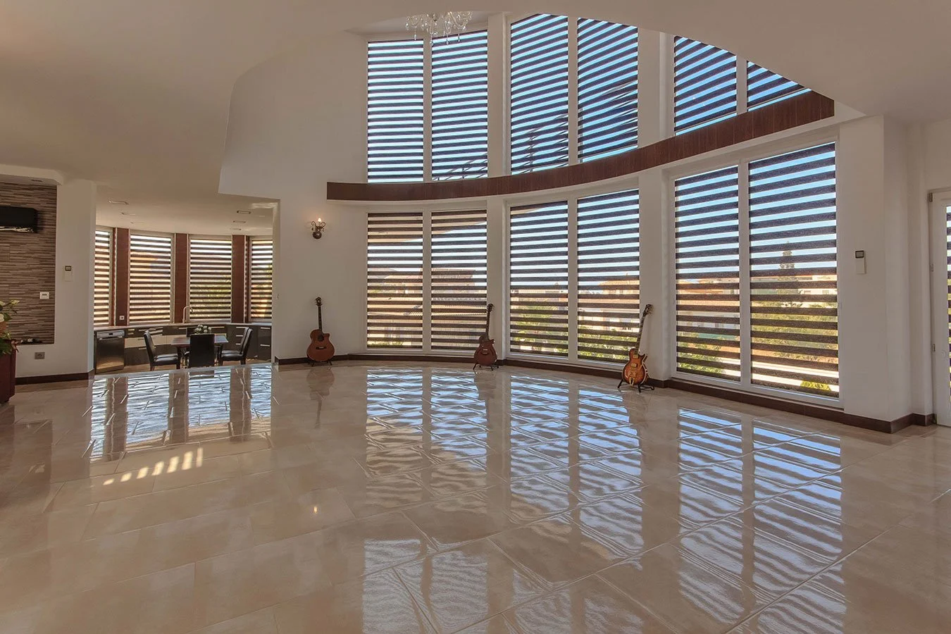 Salón amplio con ventanas grandes de persianas de listones horizontales de color oscuro, cuatro guitarras apoyadas en la pared, piso de baldosas brillante y comedor en el fondo con varias sillas.