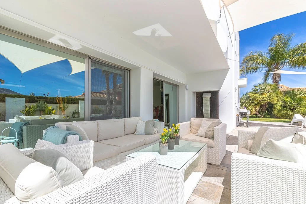 Terraza exterior con muebles de mimbre blancos y cojines, vista a un jardín con palmeras y cielo azul, en una casa moderna.