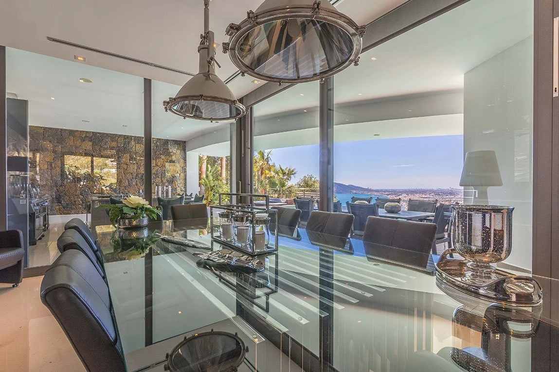 Sala de banquetes con vista panorámica al mar, mesas de vidrio, sillas negras, lámparas de techo modernas, decoración con plantas y objetos de decoración en la mesa.
