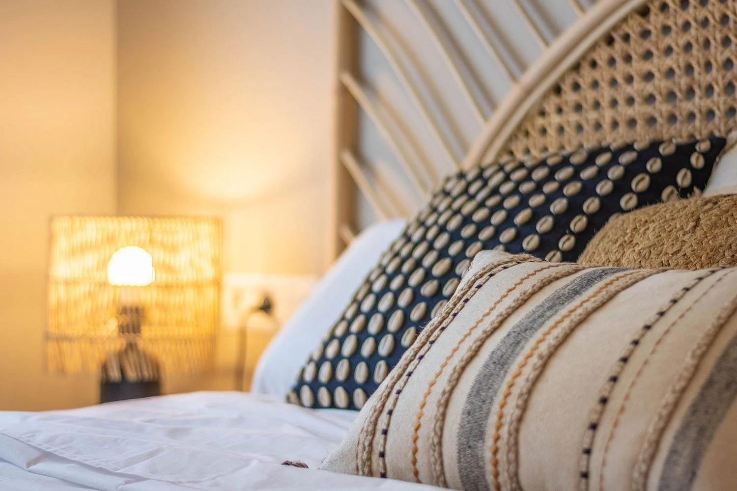 Vista cercana de varios cojines en la cama, uno de ellos con diseño de puntos blancos en fondo negro, y otro con rayas beige, gris y blancas, con cabecera de mimbre y lámpara de luz cálida en el fondo.