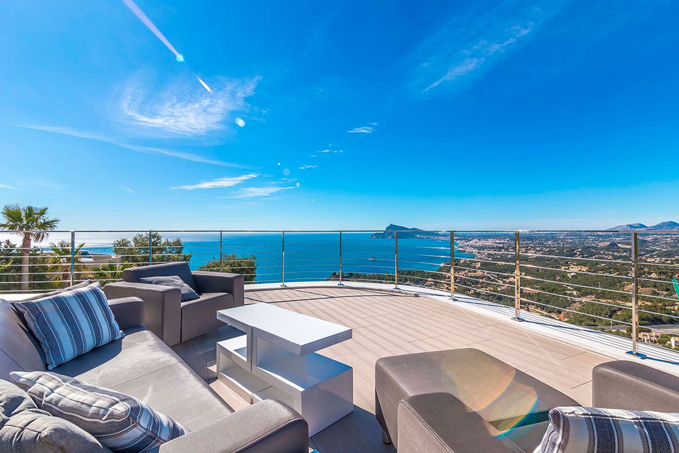 Terraza con sillones y mesa, vista panorámica del mar, la costa y montañas bajo un cielo azul con nubes y aviones