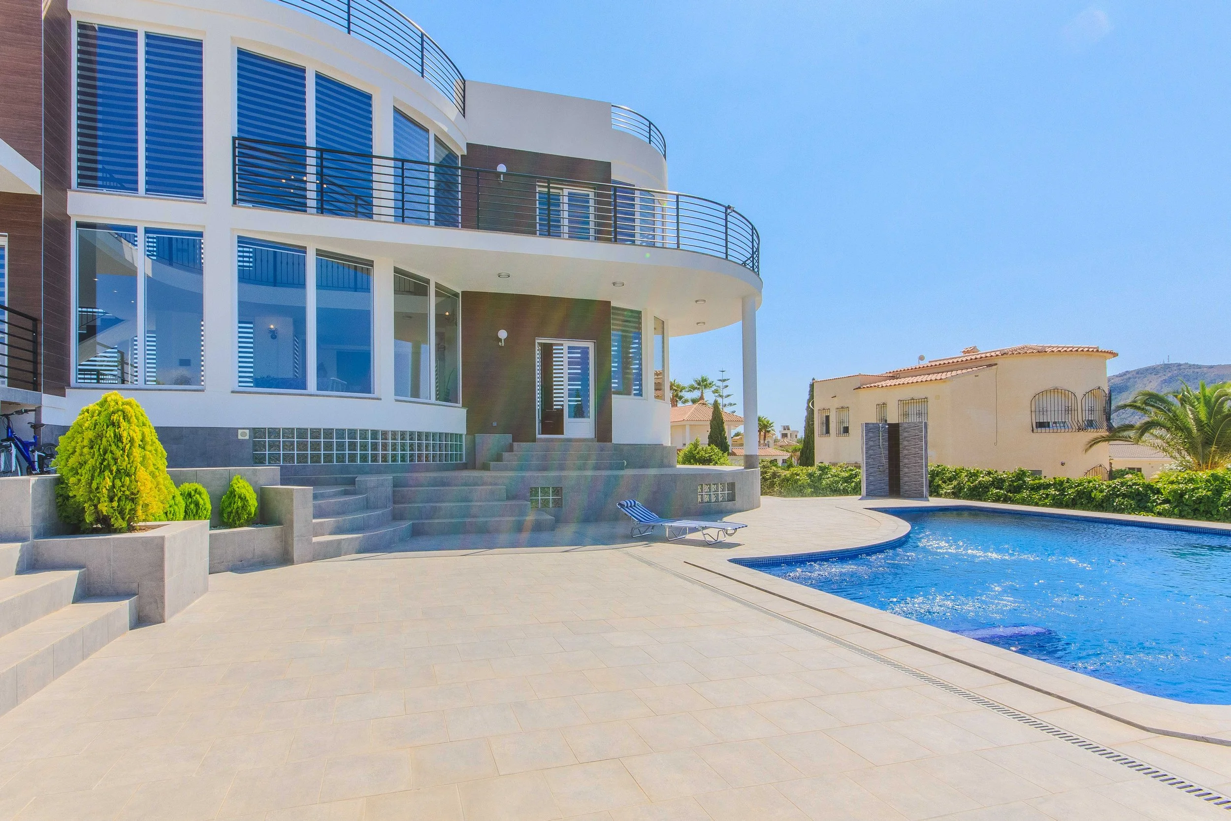 Casa moderna de varios pisos con ventanas grandes y una piscina, rodeada de un patio con plantas y una tumbona, bajo un cielo despejado y soleado.