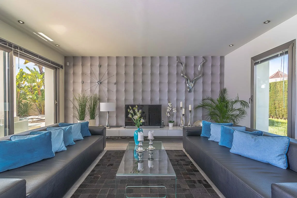 Sala de estar moderna con paredes acolchadas, sofás negros con cojines azules, mesa de cristal, plantas y decoración elegante.