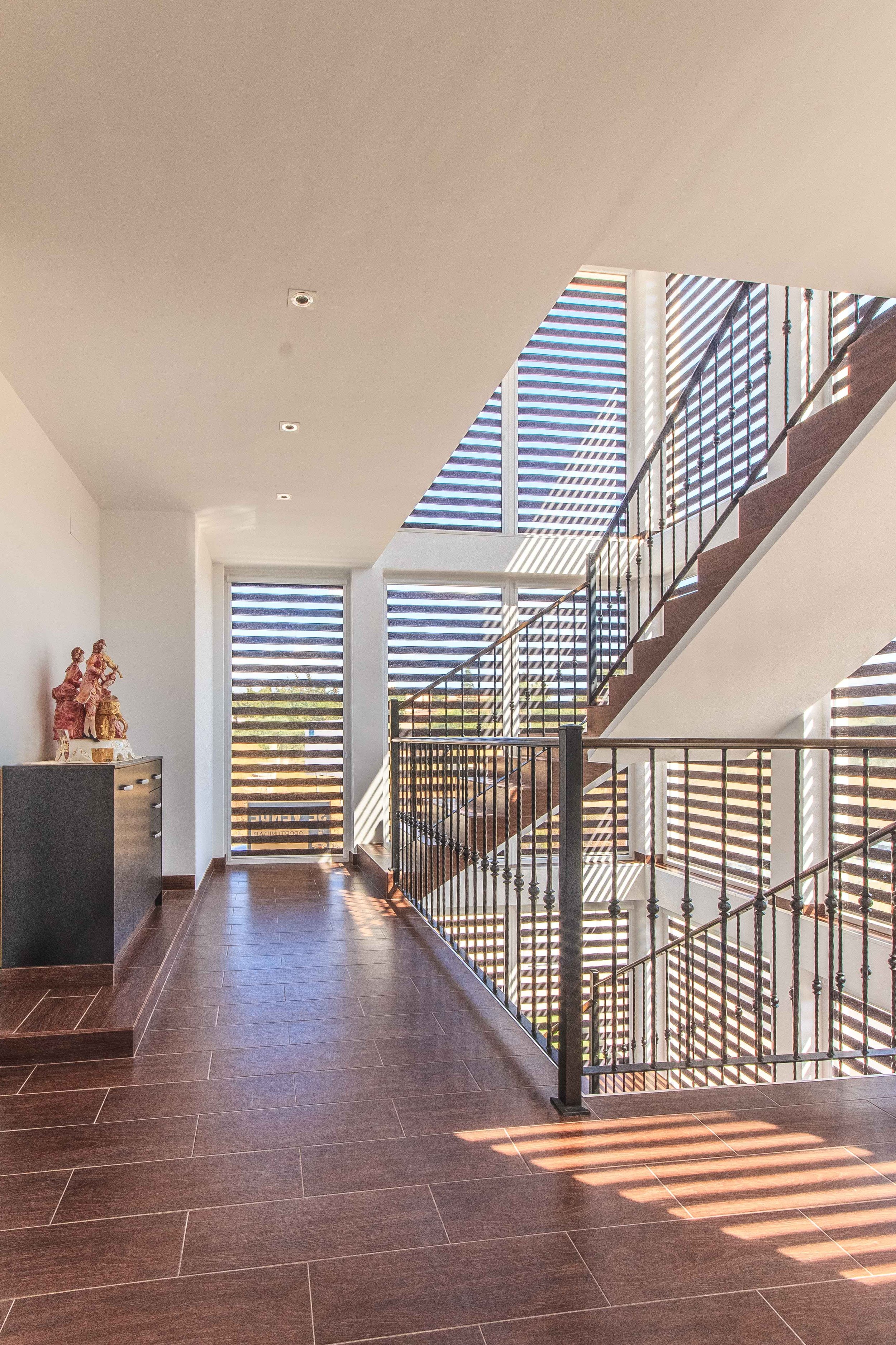 Interior de una casa moderna con escaleras de madera, barandales de metal, ventanas grandes con persianas de listones horizontales y piso de baldosas de madera, con luz natural entrando por las ventanas.