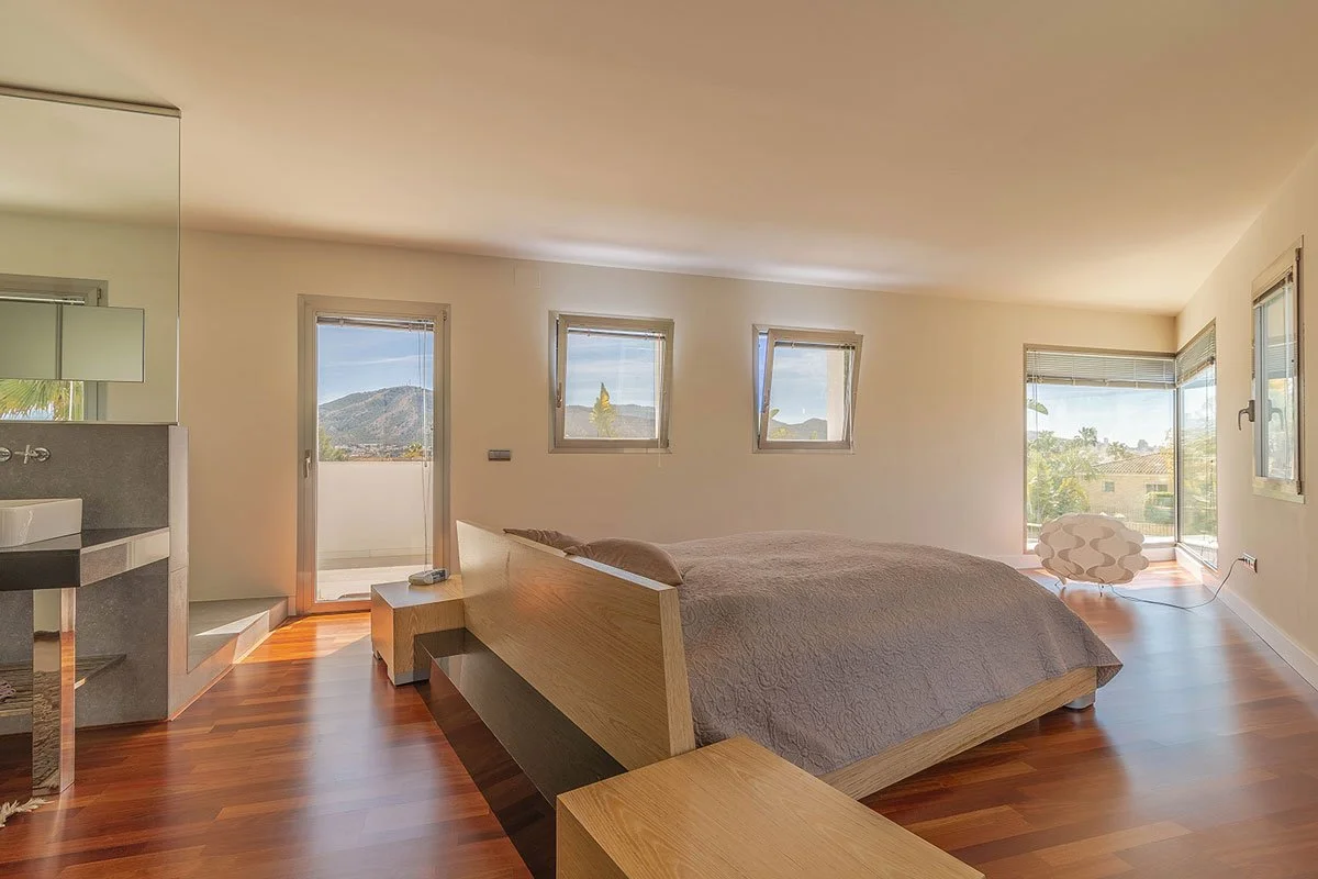 Dormitorio con ventanas y puertas de vidrio, cama doble con cabecera de madera, piso de madera, y una silla moderna junto a la ventana.