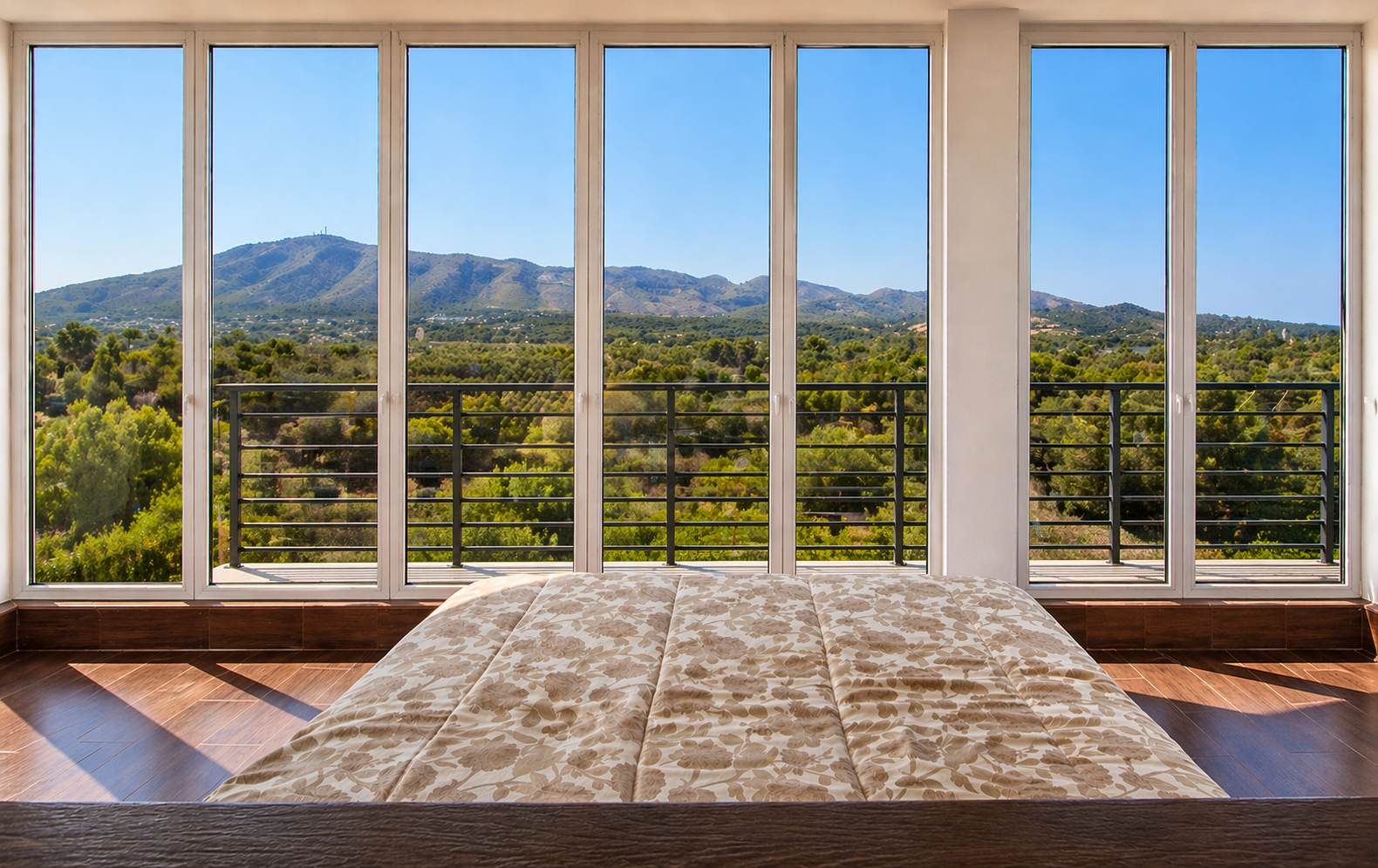 Vista de una habitación con una cama y grandes ventanas que muestran un paisaje de montañas y árboles verdes bajo un cielo despejado.