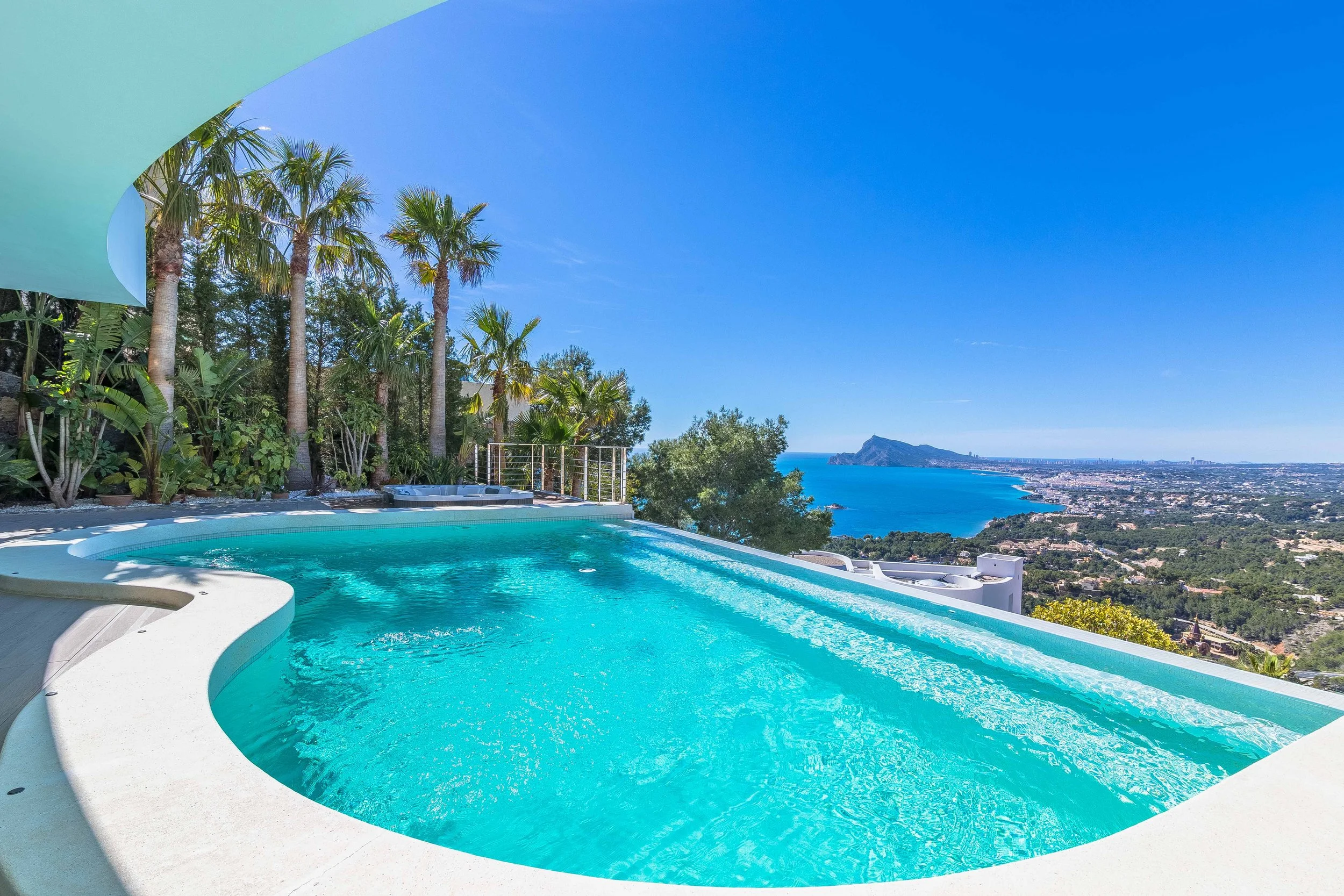 Una piscina con agua azul clara en una terraza con vista panorámica a la ciudad y al mar, rodeada de palmeras y vegetación tropical, con un cielo despejado y montañas en la distancia.