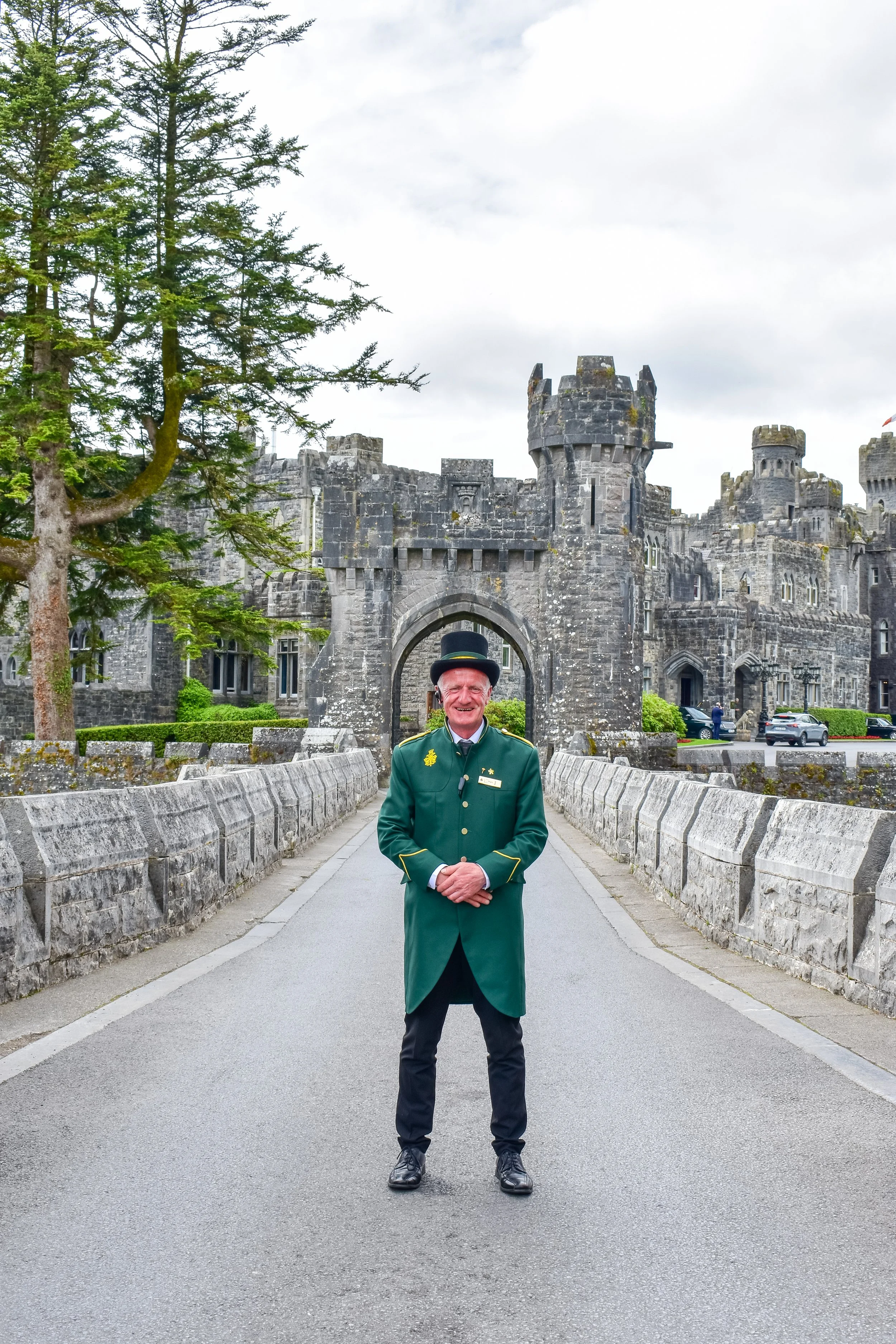 A man in a green uniform and tall black hat standing in front of a castle entrance with turrets and a stone bridge.