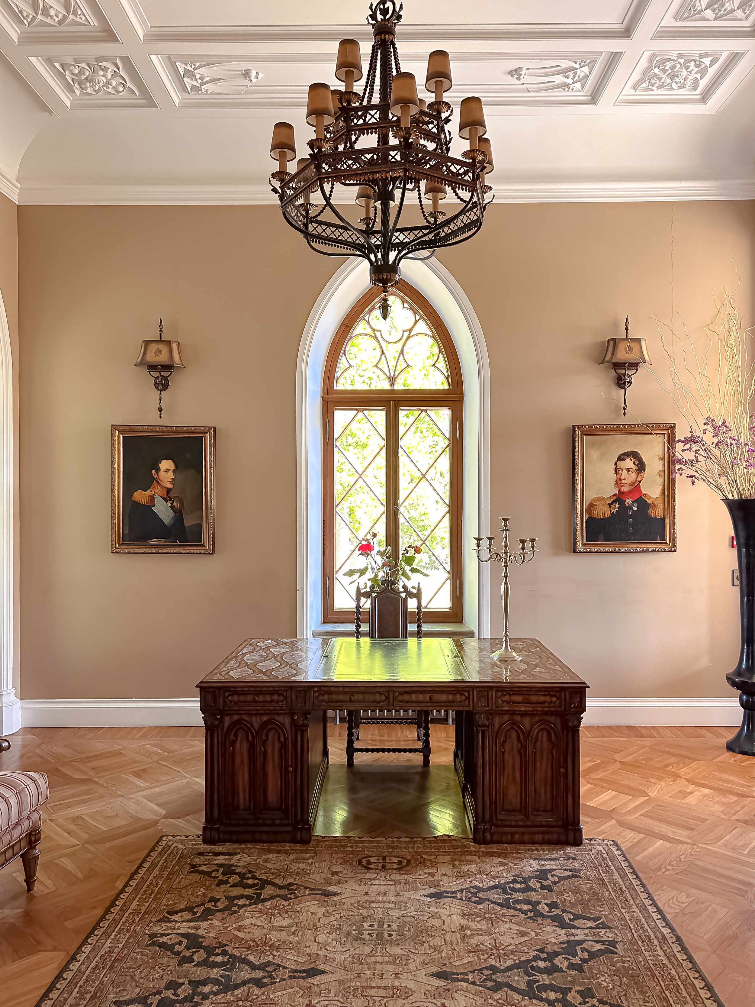 Elegant room with wooden desk, vintage chandelier, two portrait paintings, and a window with ornate design. Decor includes a candelabra, flower vase, and a patterned area rug.