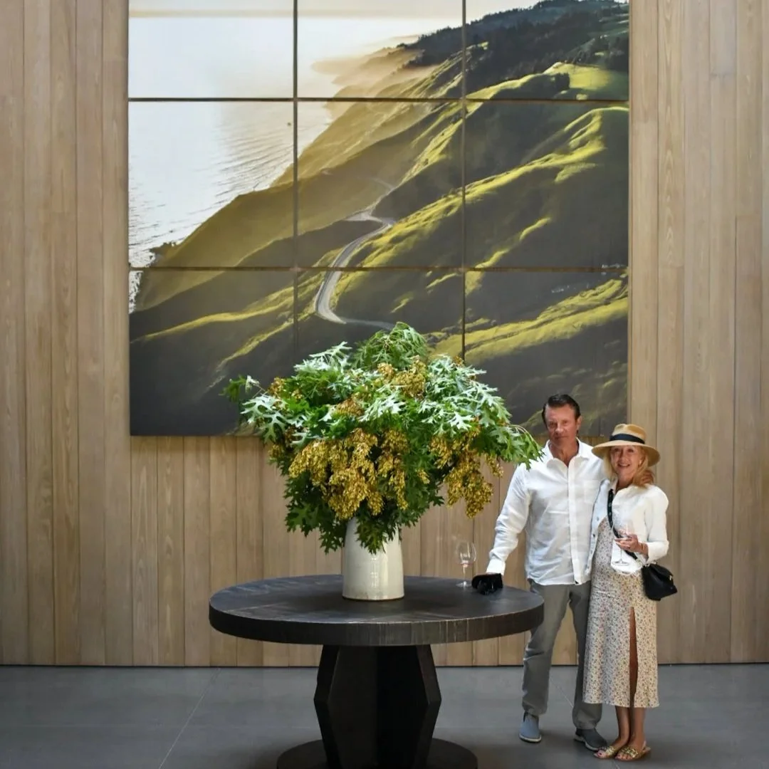 A man and woman standing next to each other in an art gallery, smiling, with a large floral arrangement on a round black table in front of them. Behind them is a large landscape photograph of rolling green hills and a winding road.