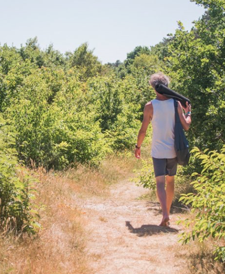 Person walking on a dirt trail surrounded by green trees and bushes, carrying a black bag over their shoulder, wearing a white sleeveless shirt and dark shorts.