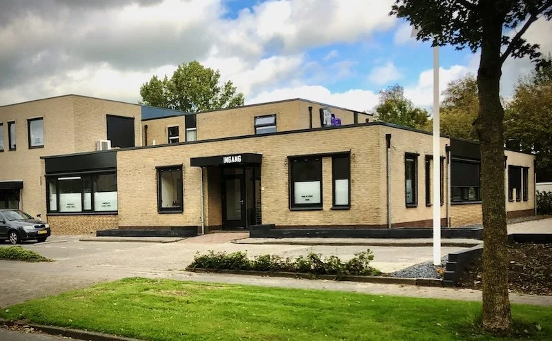 A modern brick building with a sign labeled "Ingang," surrounded by a parking lot, trees, and a small grassy area.