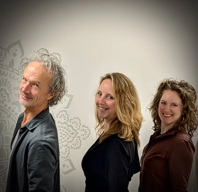 Three people, two women and one man, standing side by side, smiling at the camera in front of a plain wall with a subtle decorative pattern.