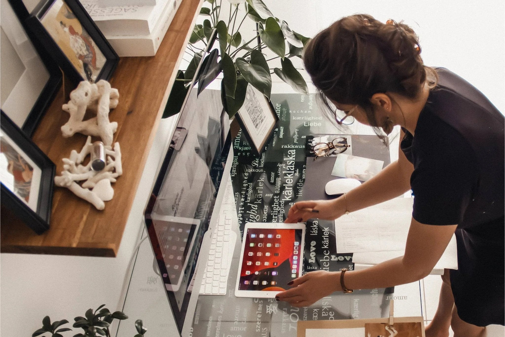 Une femme ajuste une tablette sur un bureau blanc. Le bureau est décoré avec des photos encadrées, une plante, des lunettes et des documents. Un ordinateur portable et un livre sont également présents sur le bureau.
