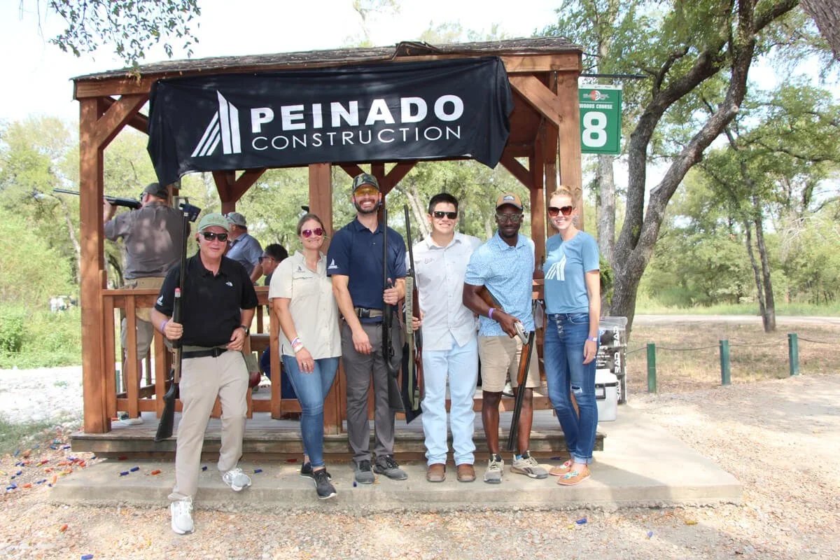Six people stand in front of a Peinado Construction sign holding shotguns