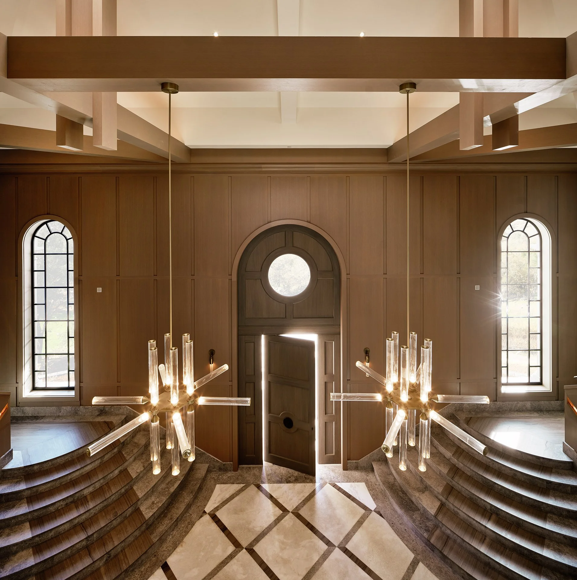 Grand foyer with curved staircases, wood-paneled walls, arched windows, starburst chandeliers, and diamond-patterned floor.