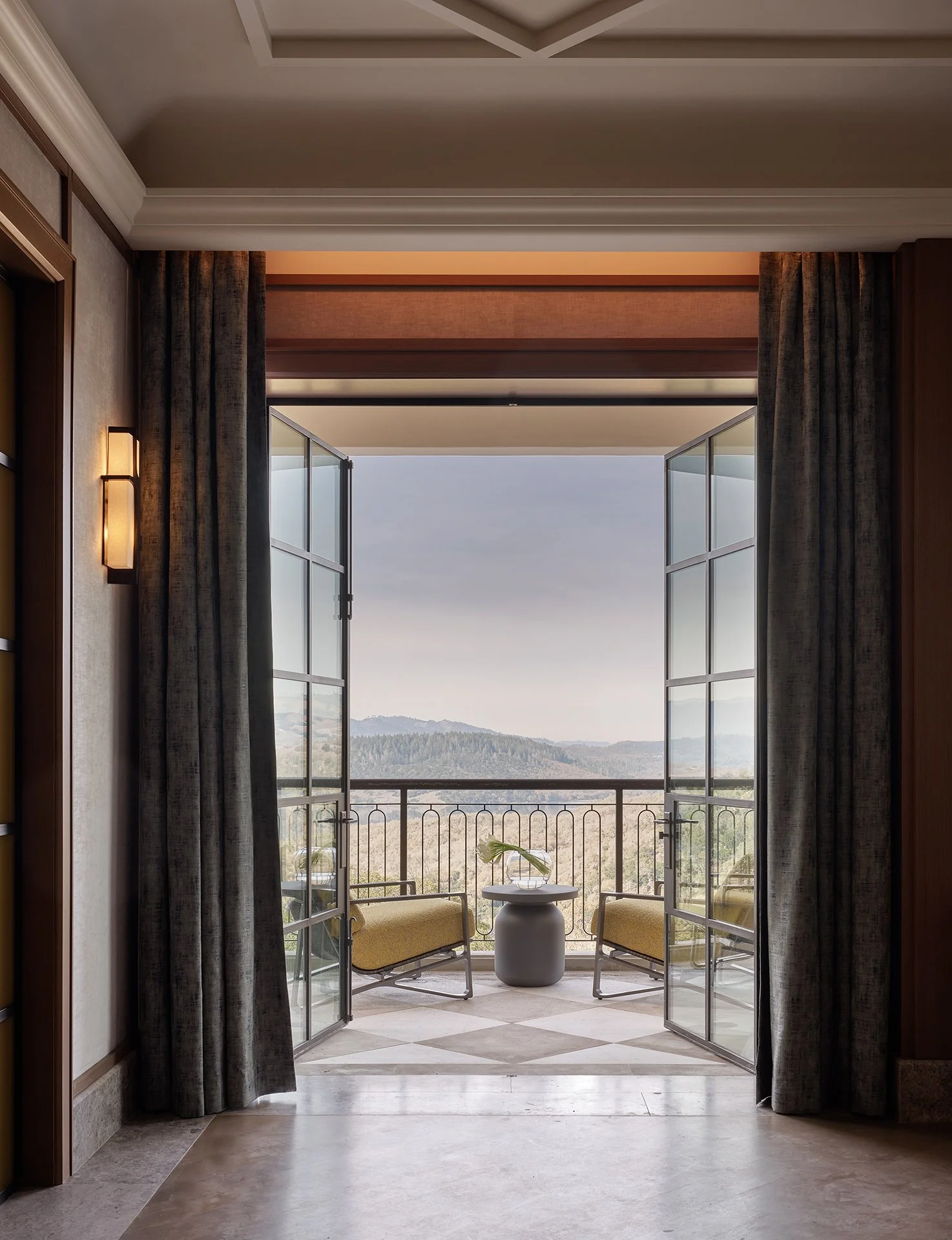 View through French doors to a private Napa terrace with yellow lounge chairs and rolling hills by Michael Wolk Design.