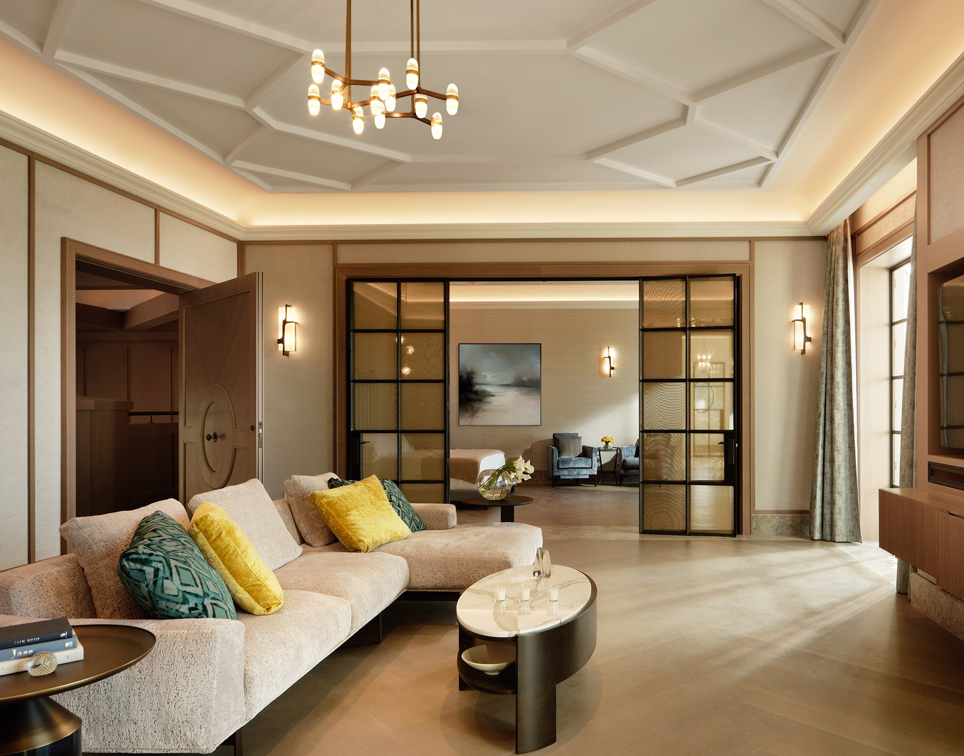 Luxurious living room with coffered ceiling, beige sectional, glass partition doors, and warm ambient lighting throughout.