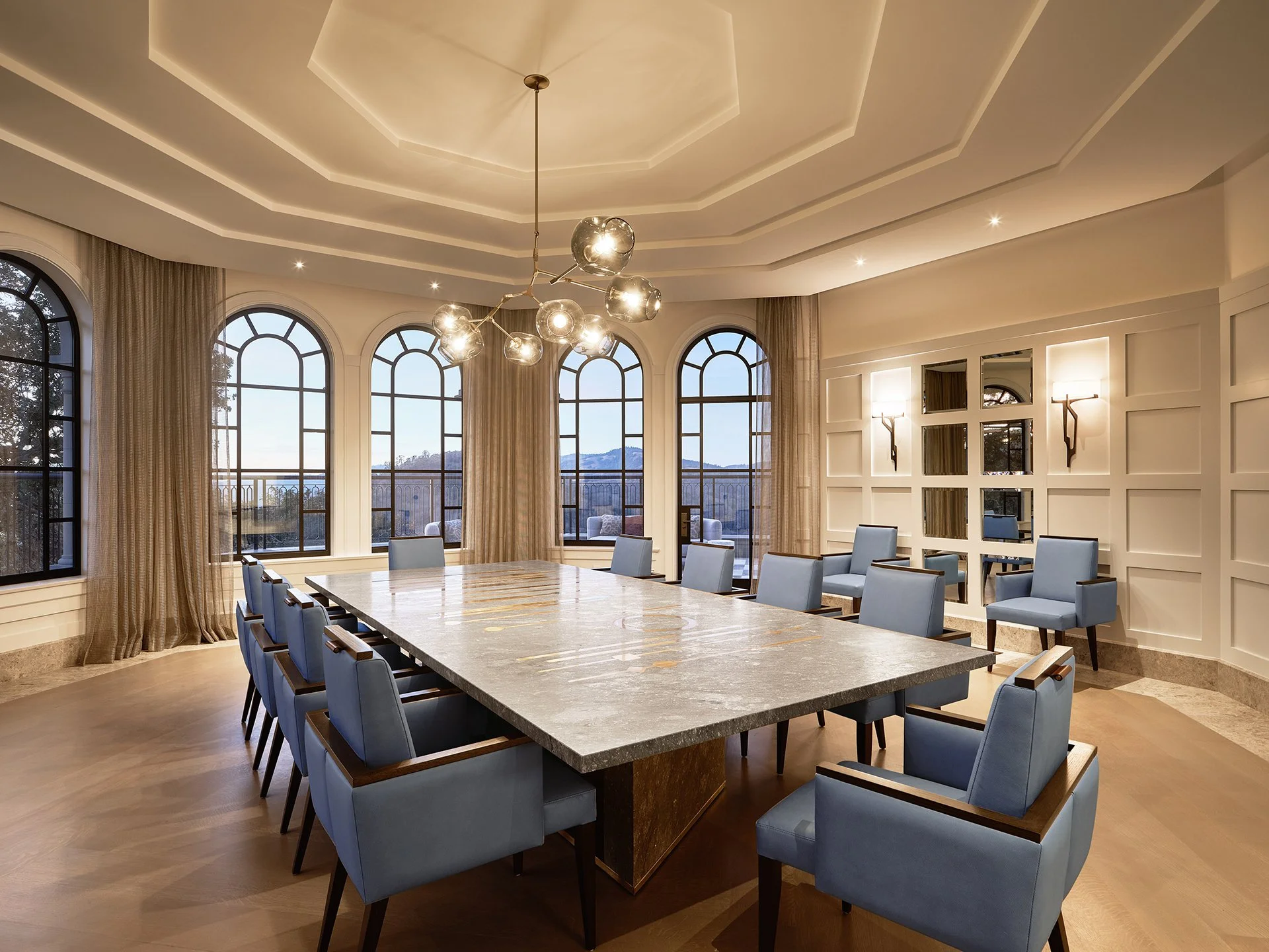 Elegant dining room: long custom stone table, modern glass chandelier, arched windows with a view, and custom upholstered chairs.