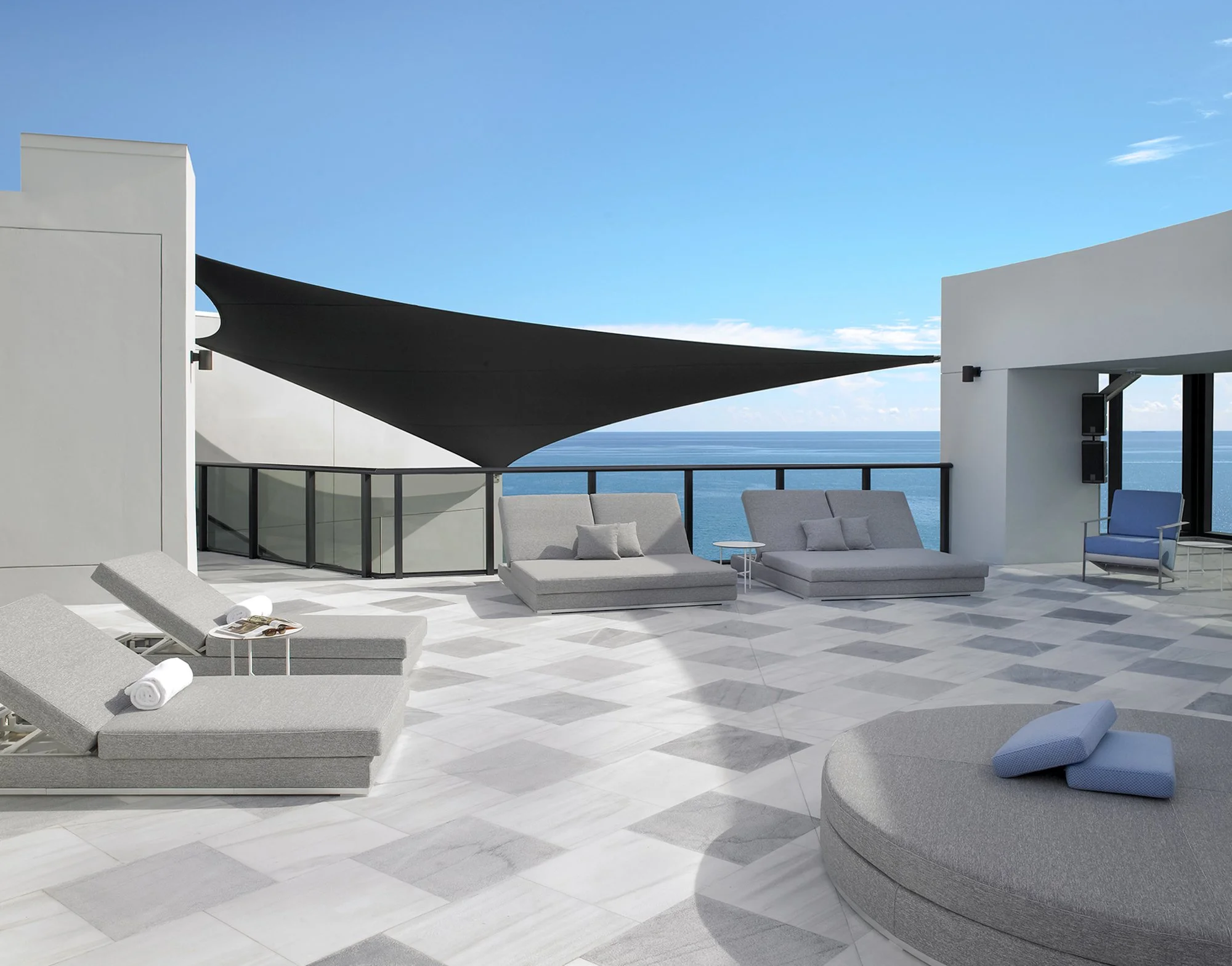 Luxury penthouse rooftop terrace with marble tile flooring, gray daybeds, shade sail, and ocean views.