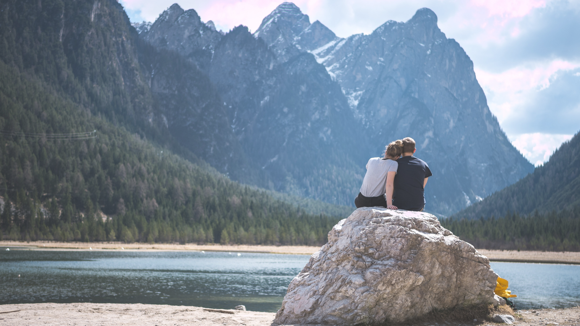 White man and woman couple sit on a rock overlooking a mountain and lake; the woman has her head on the man's shoulder talking and connecting