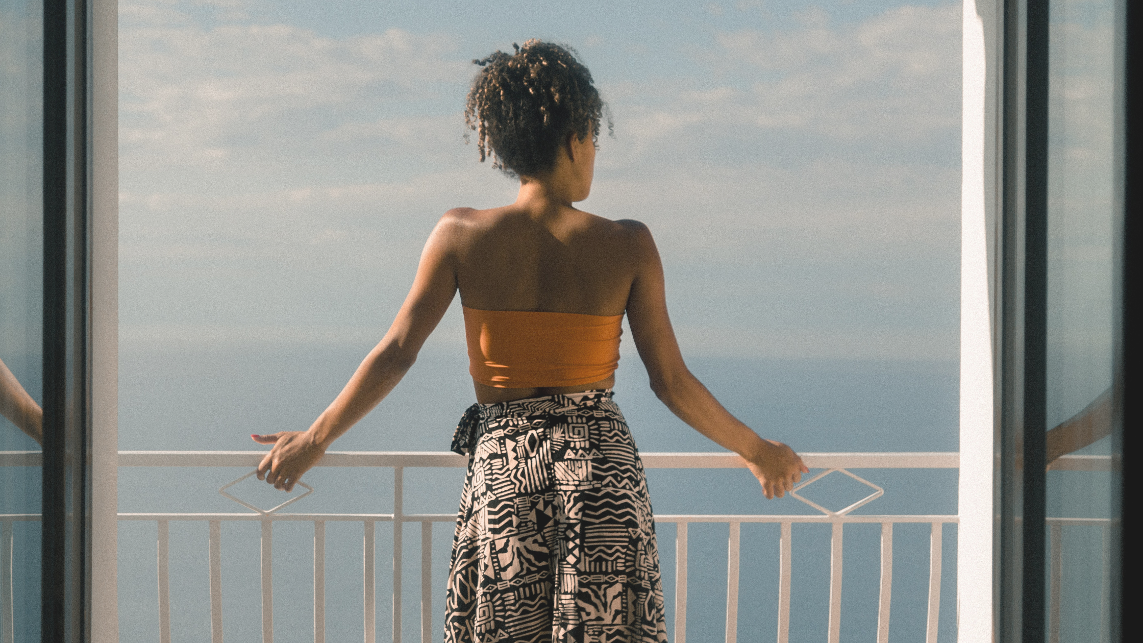 Black woman in orange crop top and black and white pattern pants stands in front of a balcony; balcony faces an open sea and blue yet cloudy sky