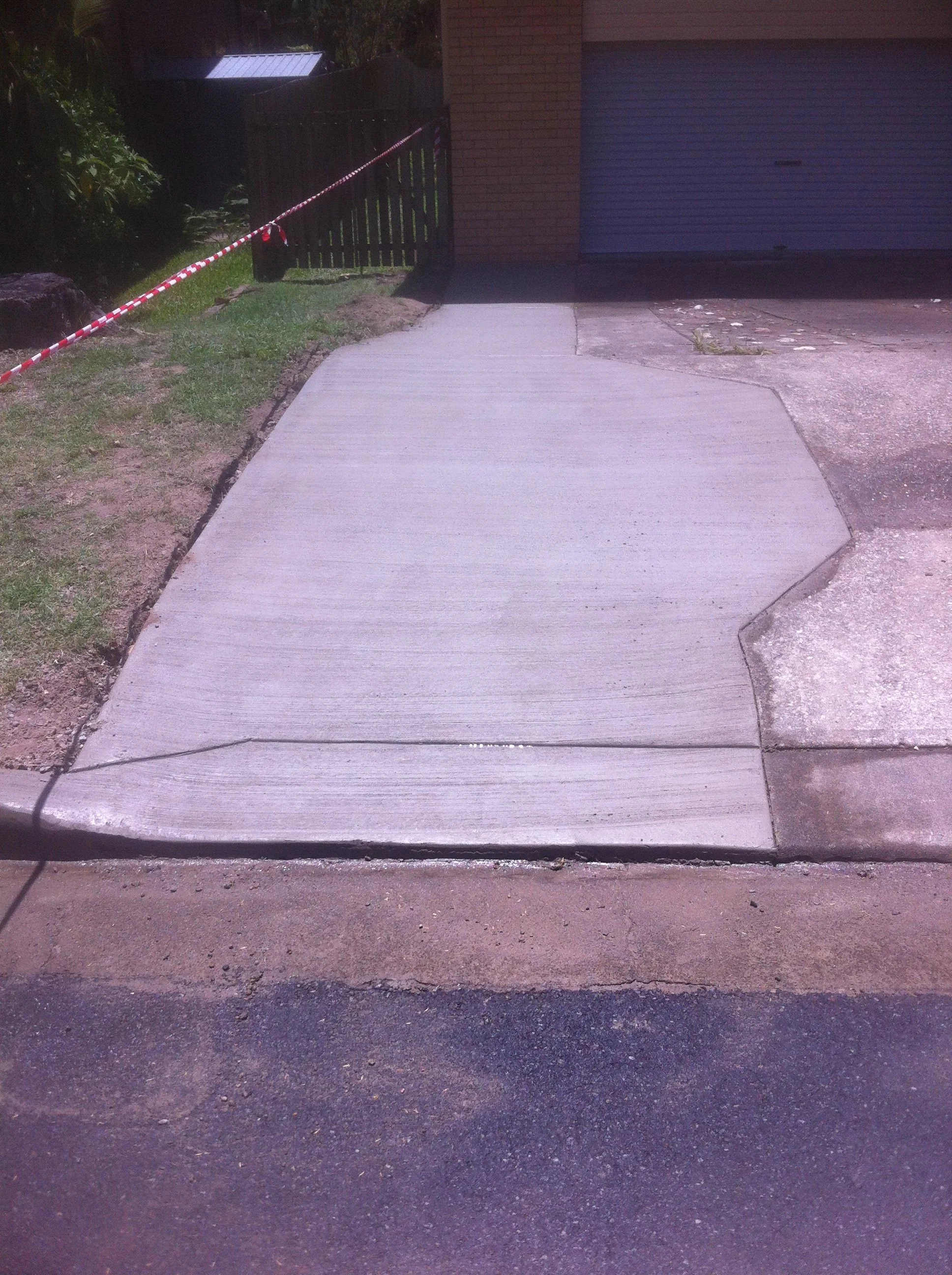 Freshly poured concrete sidewalk beside a brick garage, bordered by red-and-white caution tape.