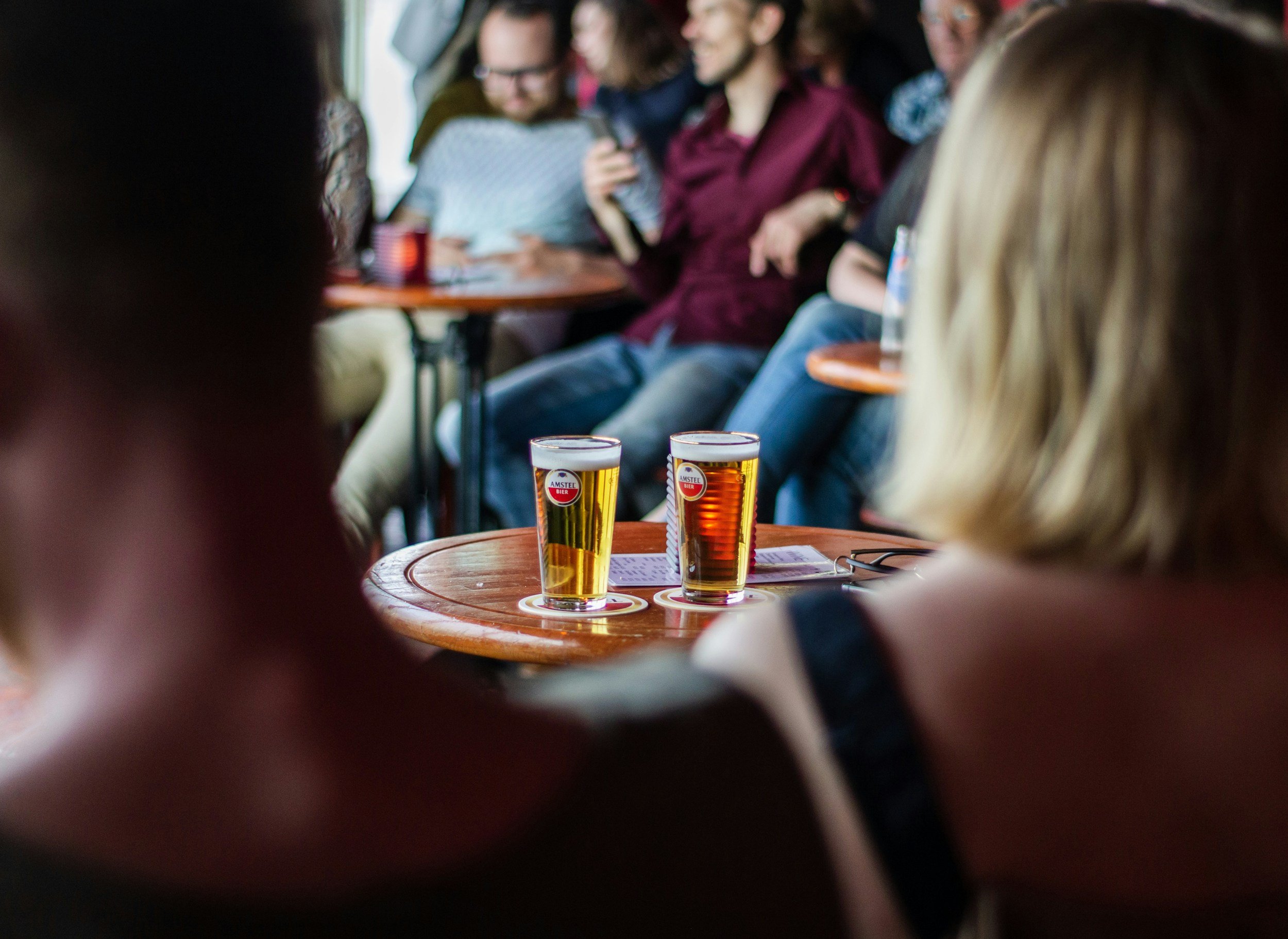Three glasses of beer with labels on a wooden table in a social setting, blurred background of people talking and smiling.