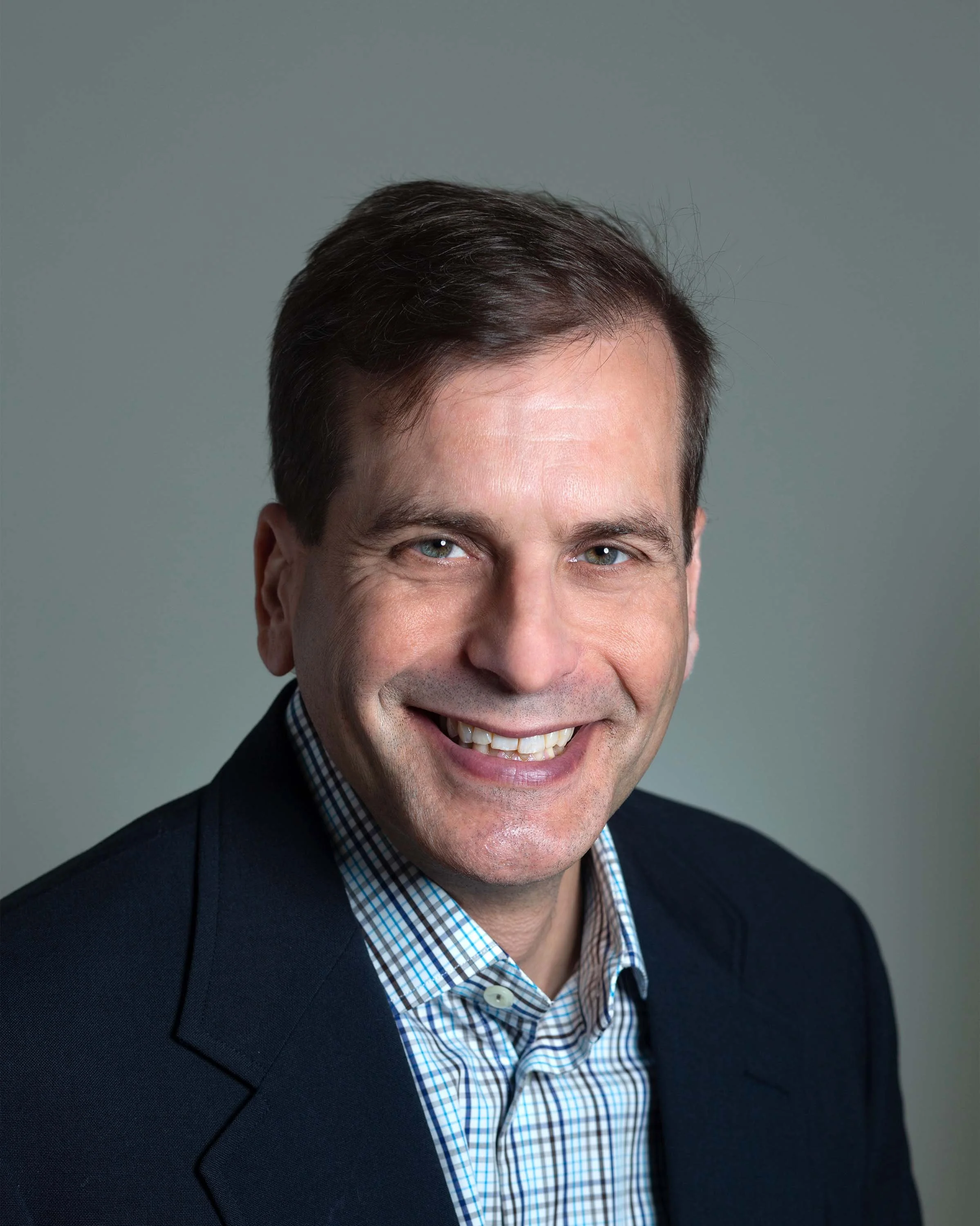 Professional portrait of a smiling man with dark hair, wearing a dark blazer and a checkered shirt, against a plain background.