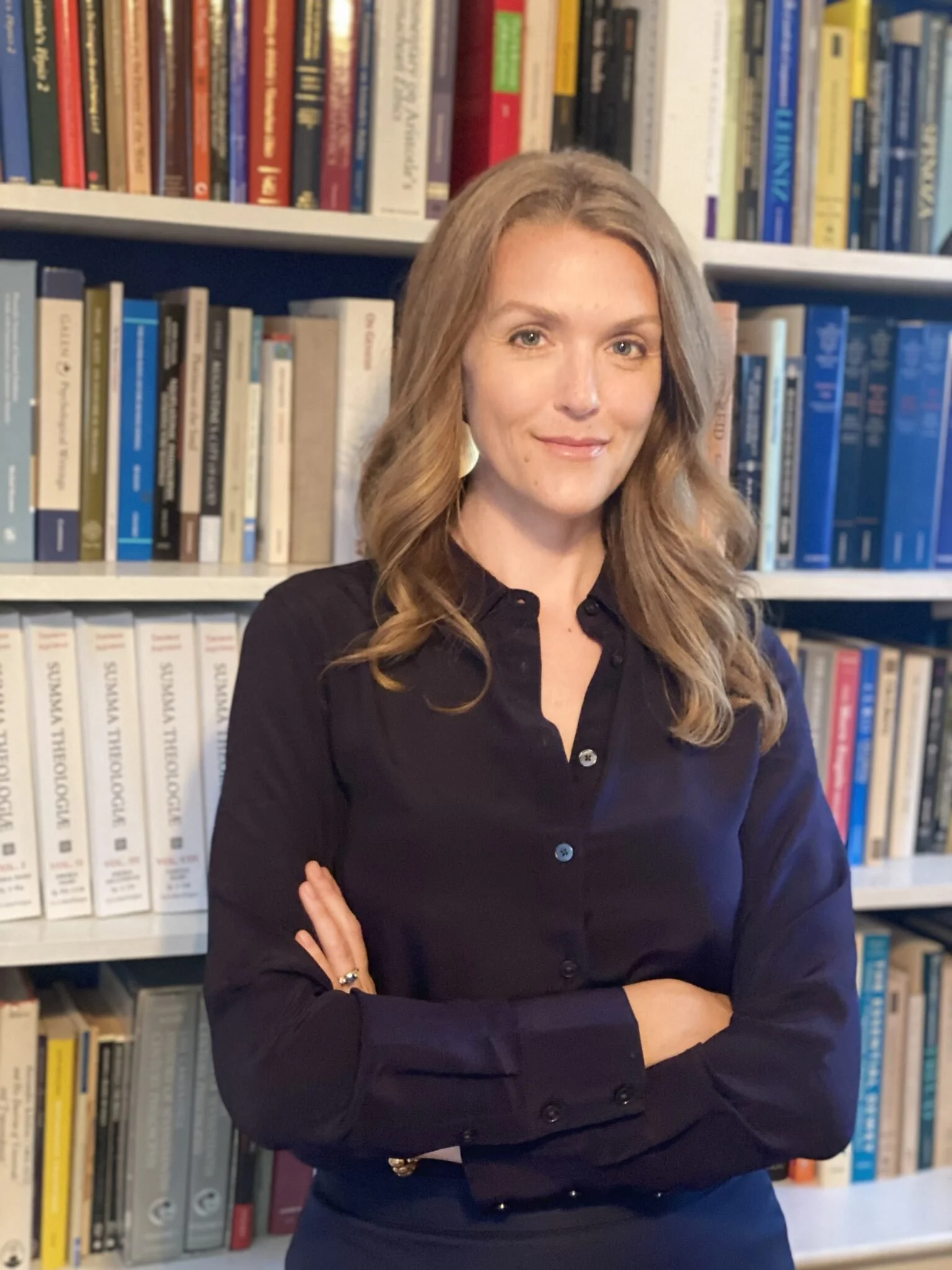 A woman with wavy blonde hair and light skin standing with arms crossed in front of bookshelves filled with various books.