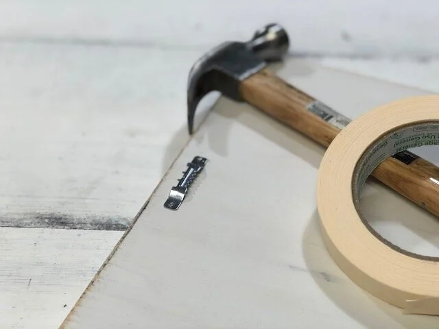 How to attach a sawtooth hanger to a wood sign. Quick and easy way to put hanging hardware on a sign to hang. Use masking tape to hold your sawtooth hanger in place before you hammer it down.