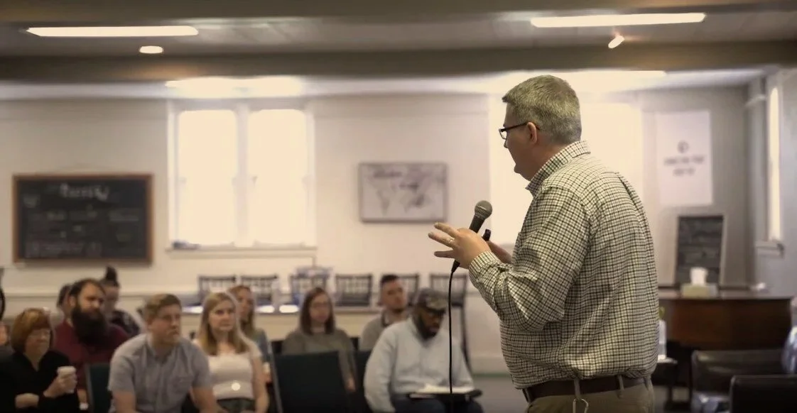A man with a microphone speaking to a small seated crowd.