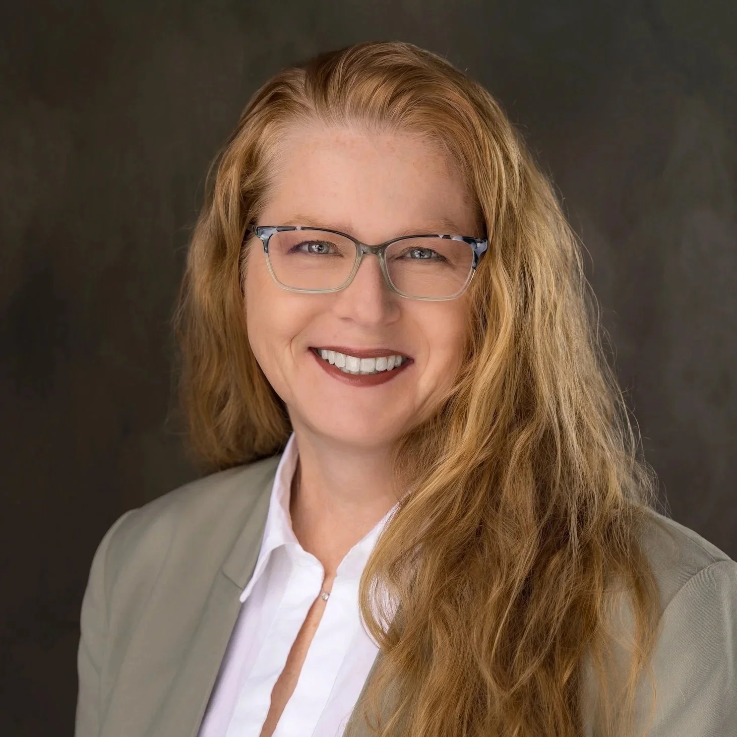 A professional woman with long red hair, glasses, and a smile, wearing a light blazer over a white shirt, posing against a dark background.