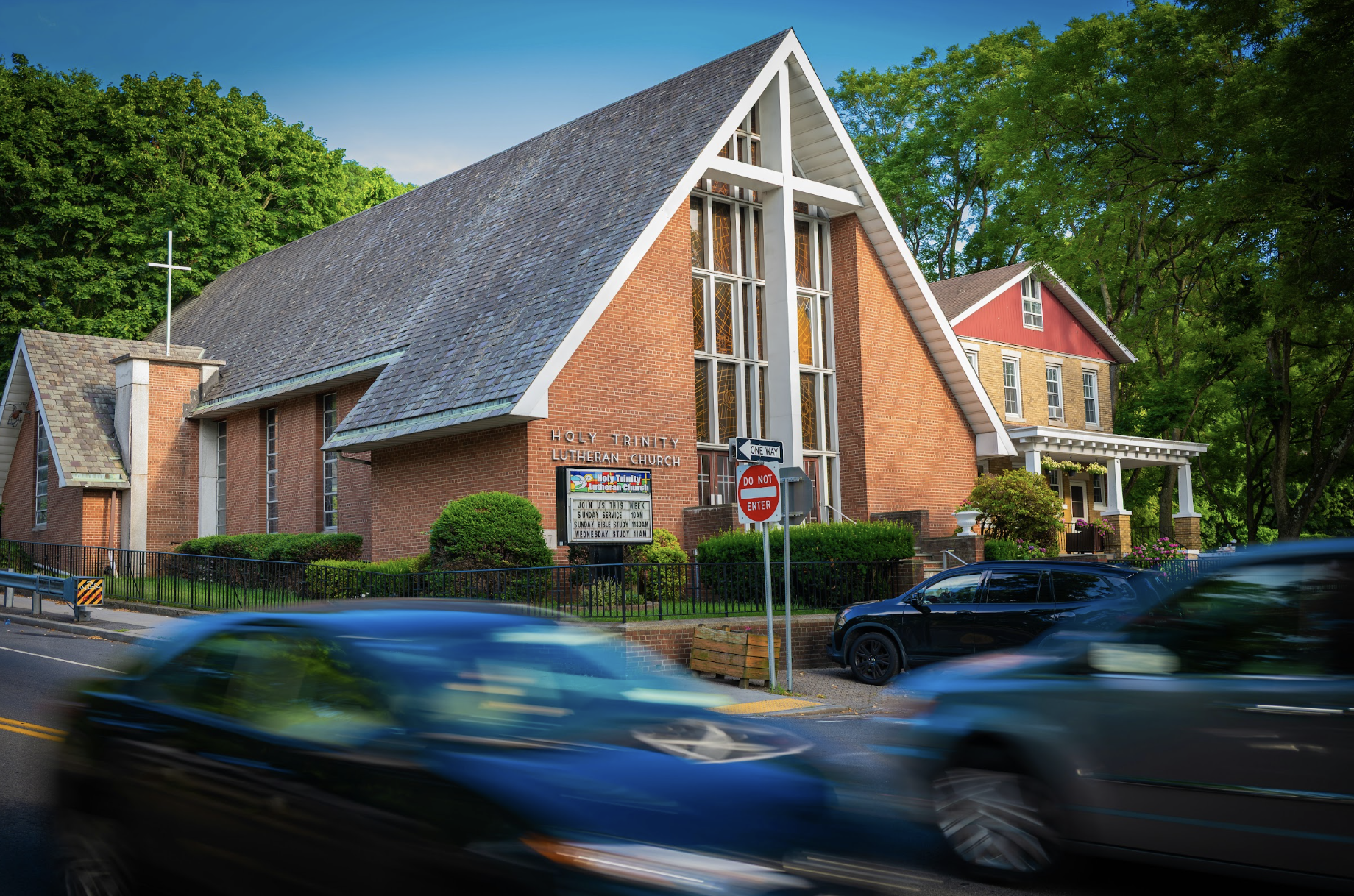 Exterior view of a Lutheran church, featuring a red brick facade with a large white cross emblem, as cars pass by on the street in front.