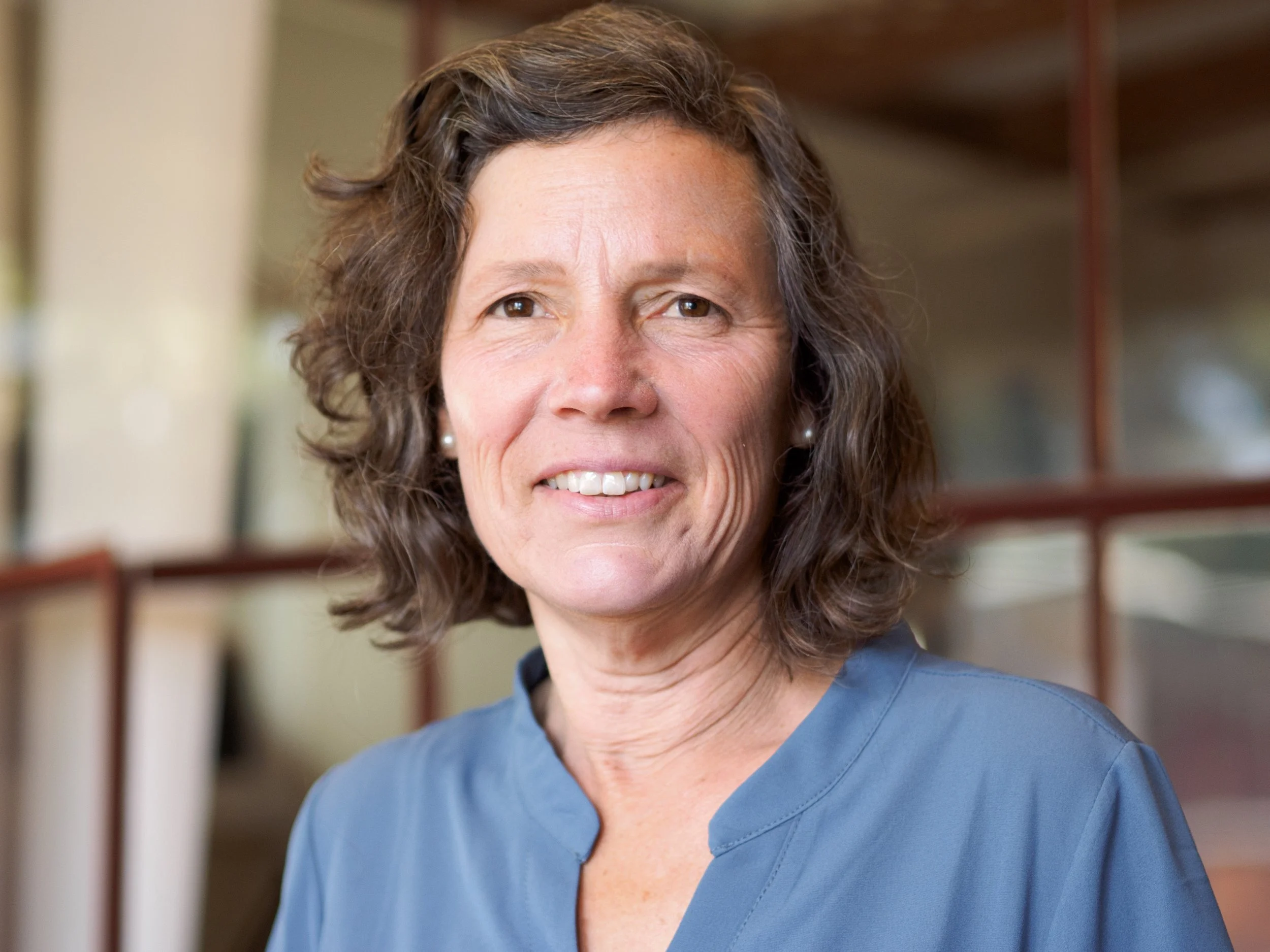 A portrait of a woman with short wavy brown and gray hair in a blue blouse