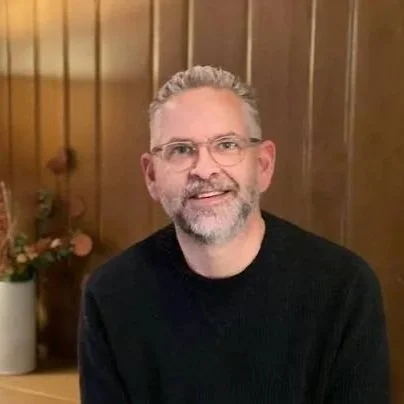 A portrait of a man with gray hair, a gray beard, glasses, and a black sweater sitting in front of a wood paneled wall