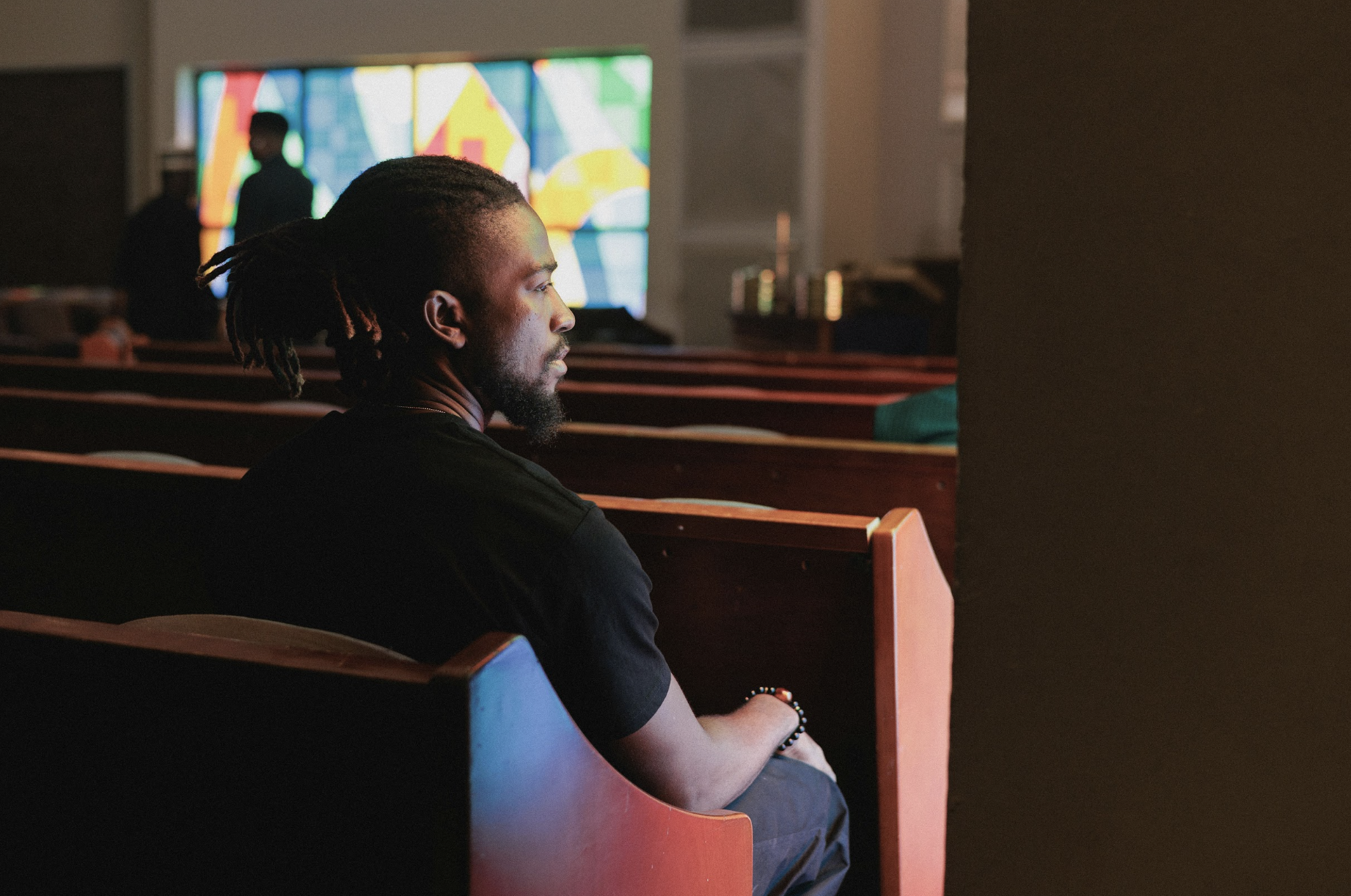 A man with dreadlocks tied back sits alone in a church pew, looking towards a colorful stained-glass window in the background.