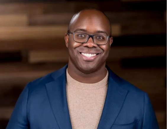 A smiling bald man with black glasses in a beige sweater and blue sports coat in front of wood paneled wall