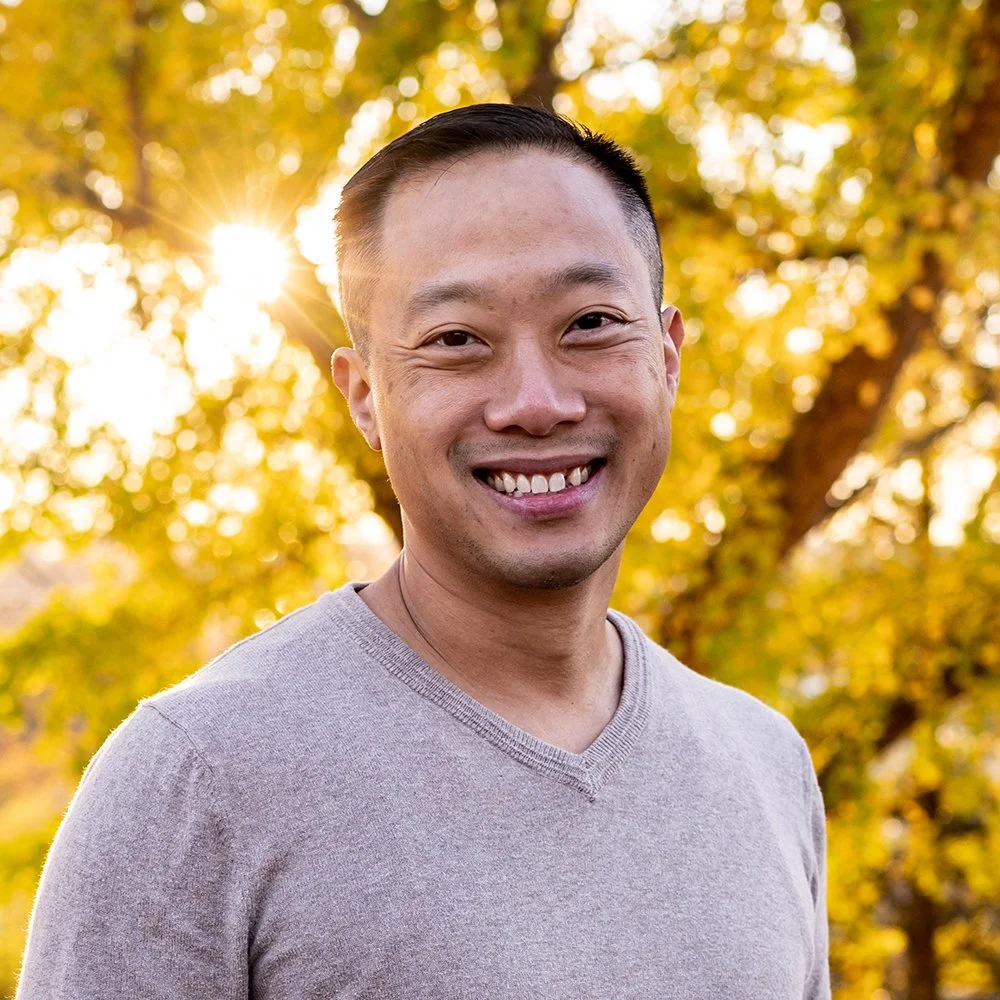 A smiling young man with short black hair in a grey t-shirt standing outside in the fall sunshine