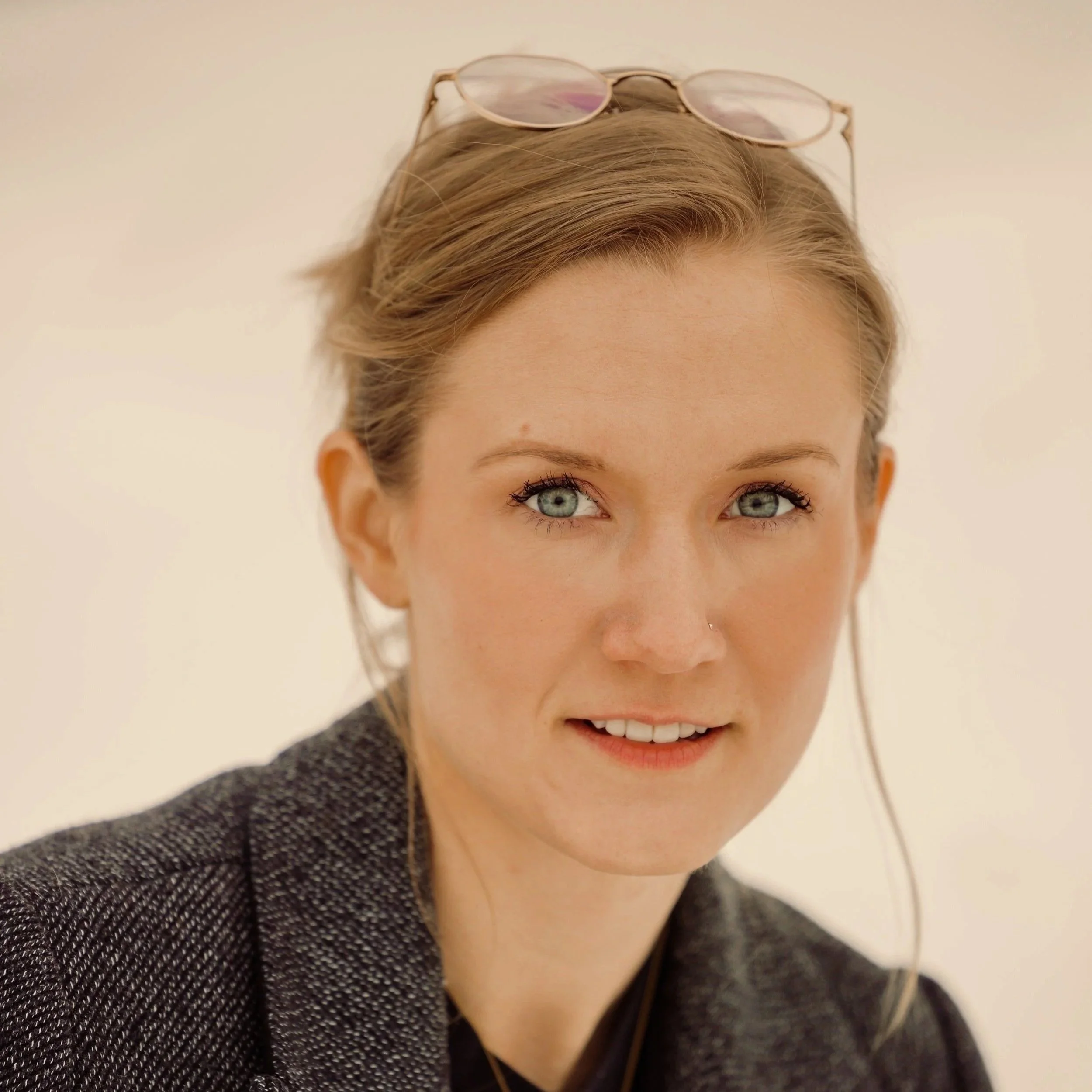 Portrait of a young woman with blonde hair pulled up in a clip, glasses on her head, and wearing a tweed jacket