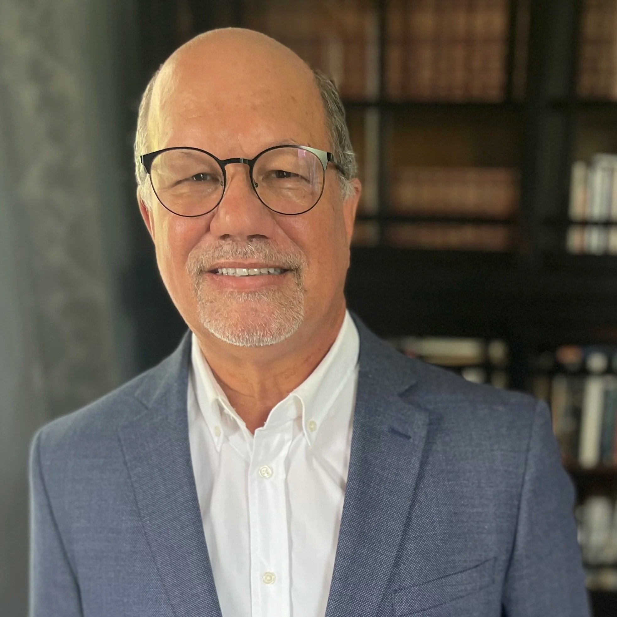 A man in glasses with a gray beard in a blue suit jacket and white shirt with bookshelves in the background.