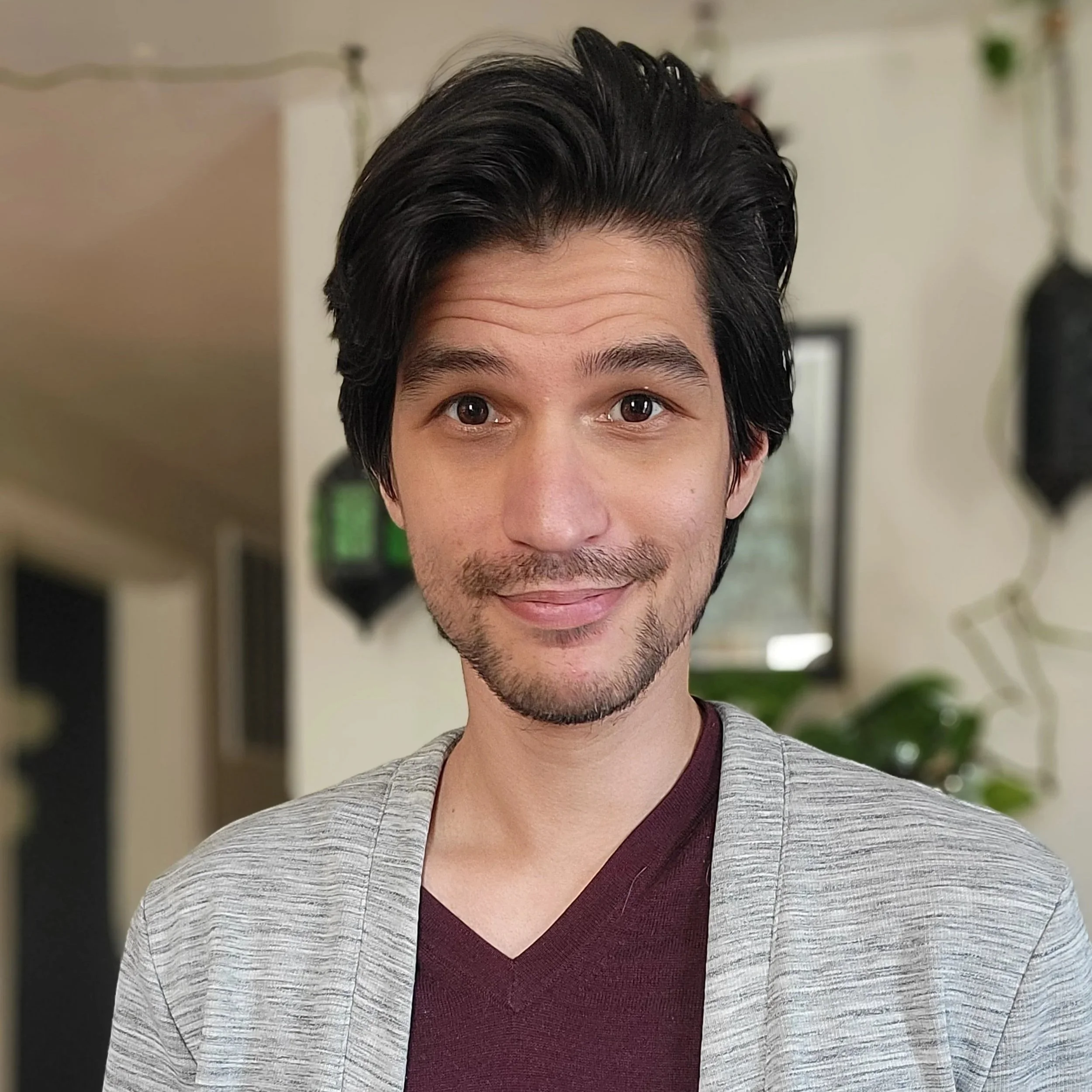 A smiling young man with dark hair and short dark facial hair in a grey cardigan and maroon t shirt
