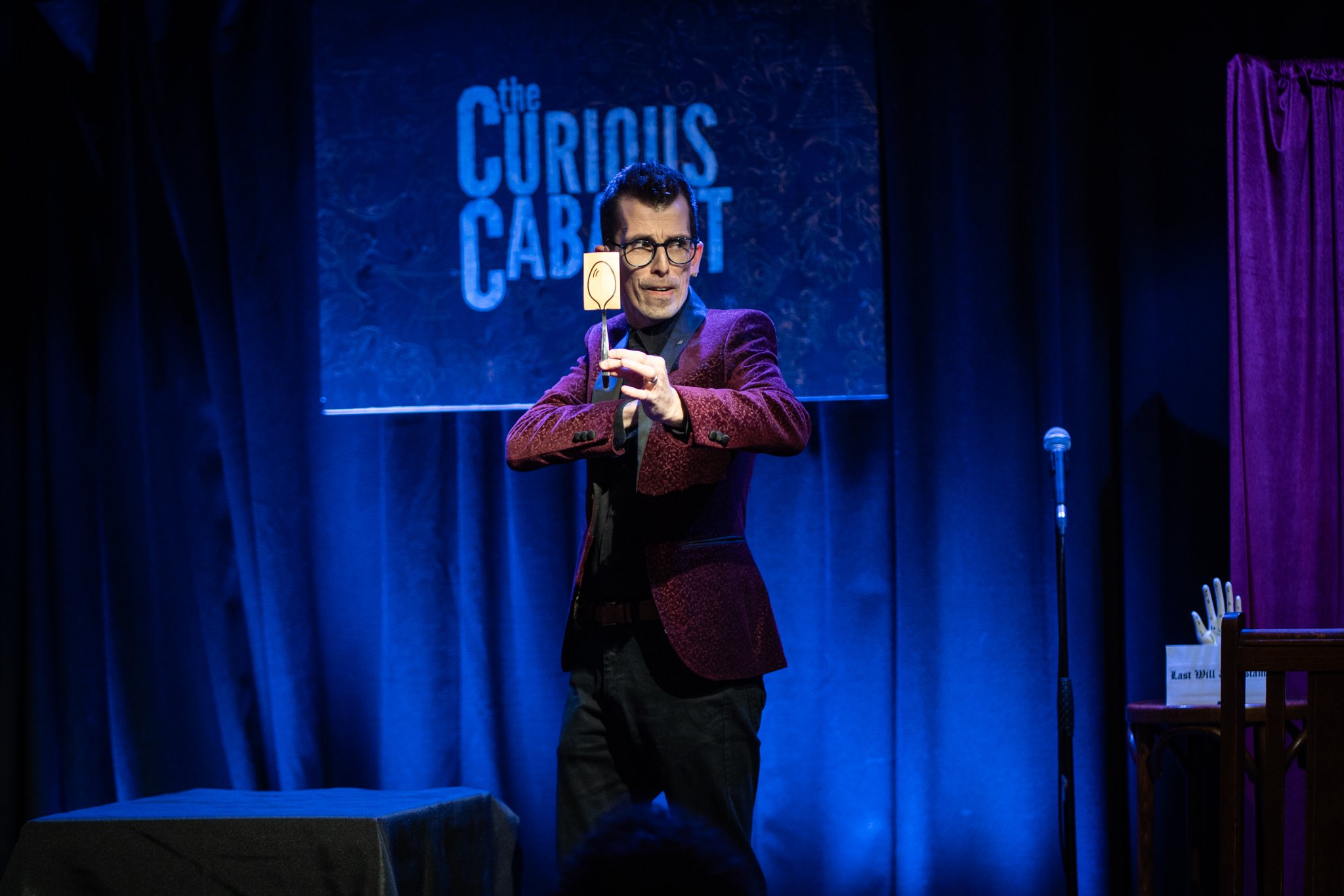 A man wearing glasses and a maroon blazer performs on stage at a comedy show titled 'The Curious Cabinet', holding a prop in the shape of an ear.