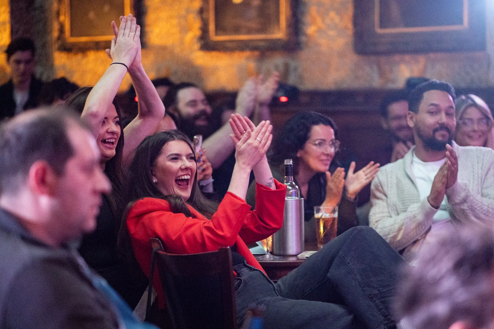 People sitting in a dimly lit bar, clapping and laughing during a comedy show or performance.