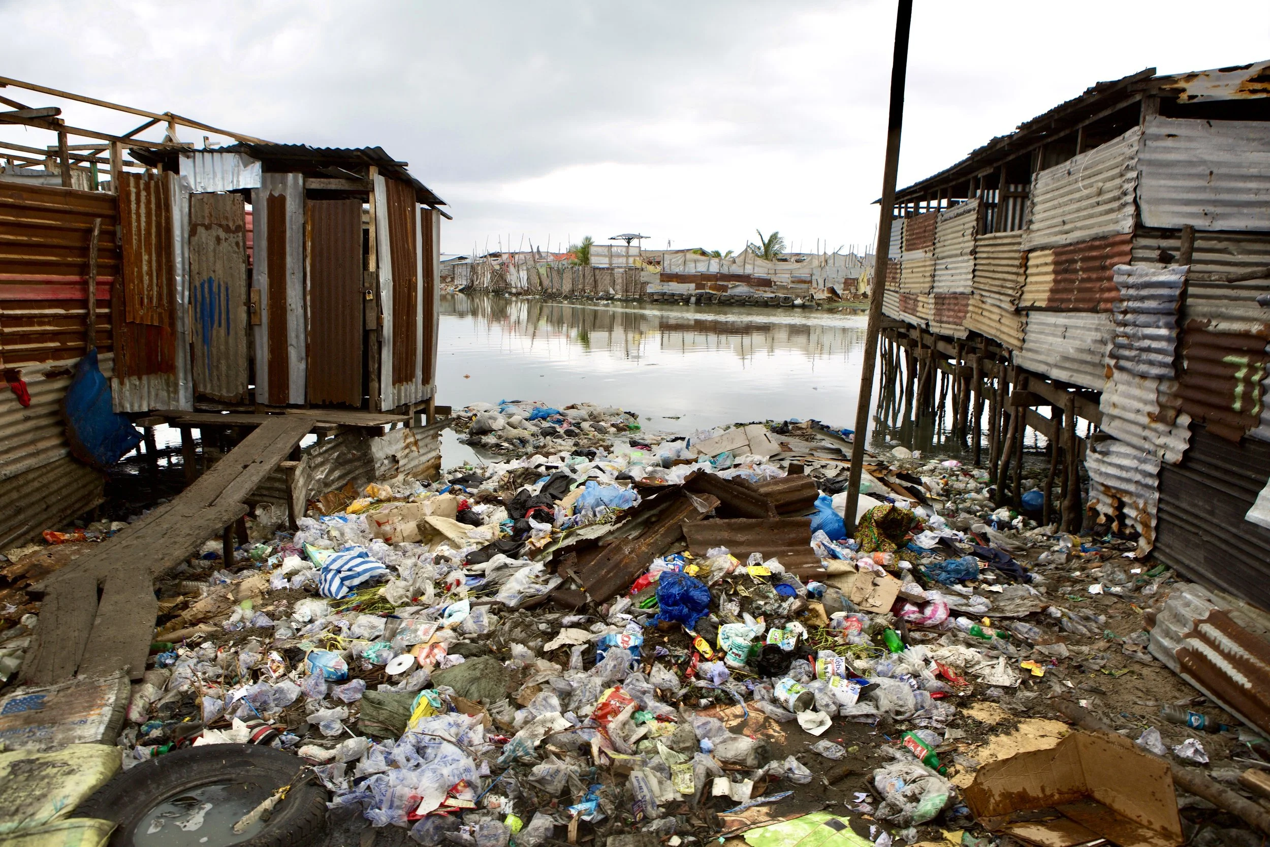 a flooded area with makeshift houses made of corrugated metal and wood, surrounded by trash and debris, with water covering the ground and reflecting the structures