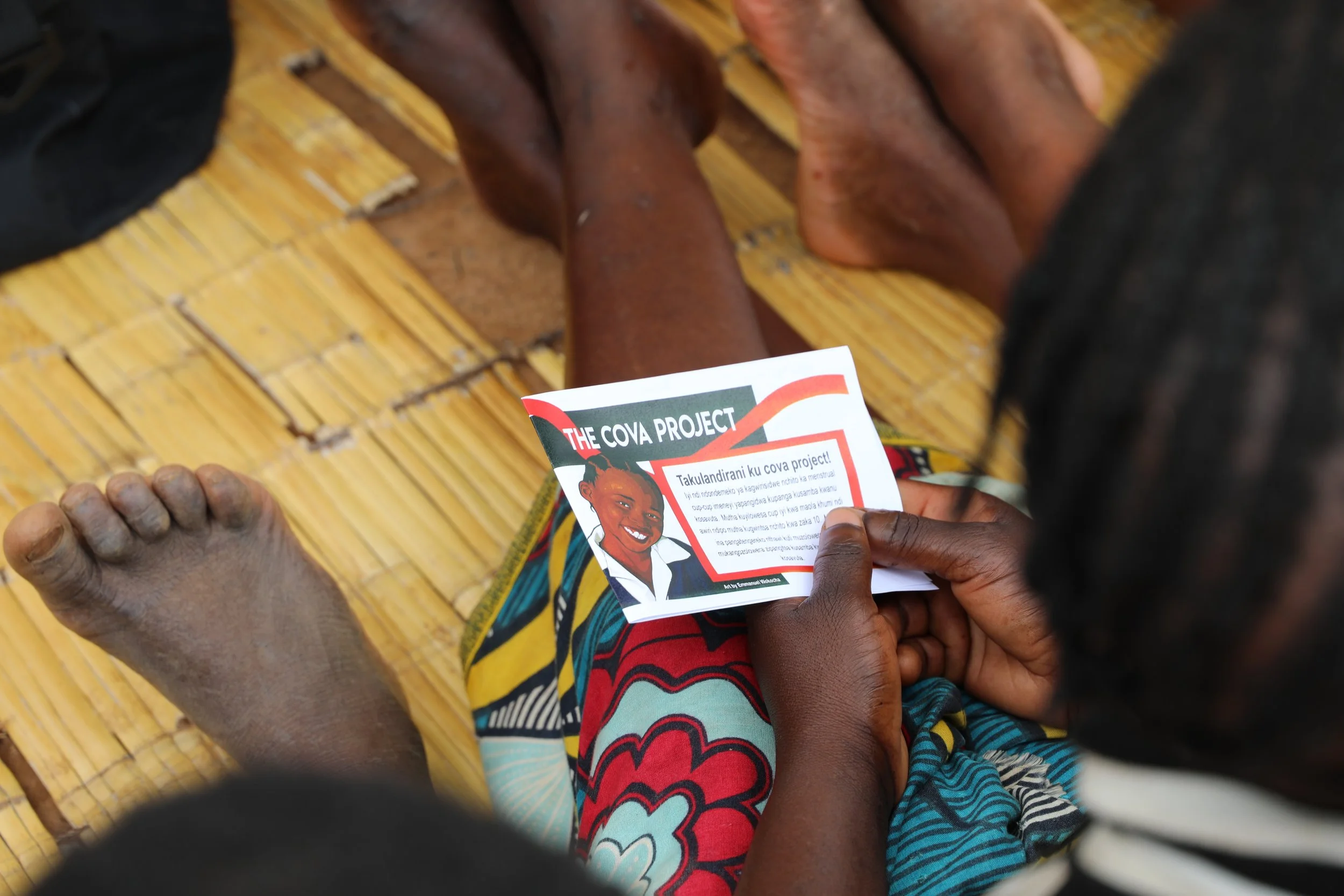 Close-up of a person holding a flyer or pamphlet with a smiling woman on it, with a focus on the hands and flyer, sitting on a woven bamboo mat.